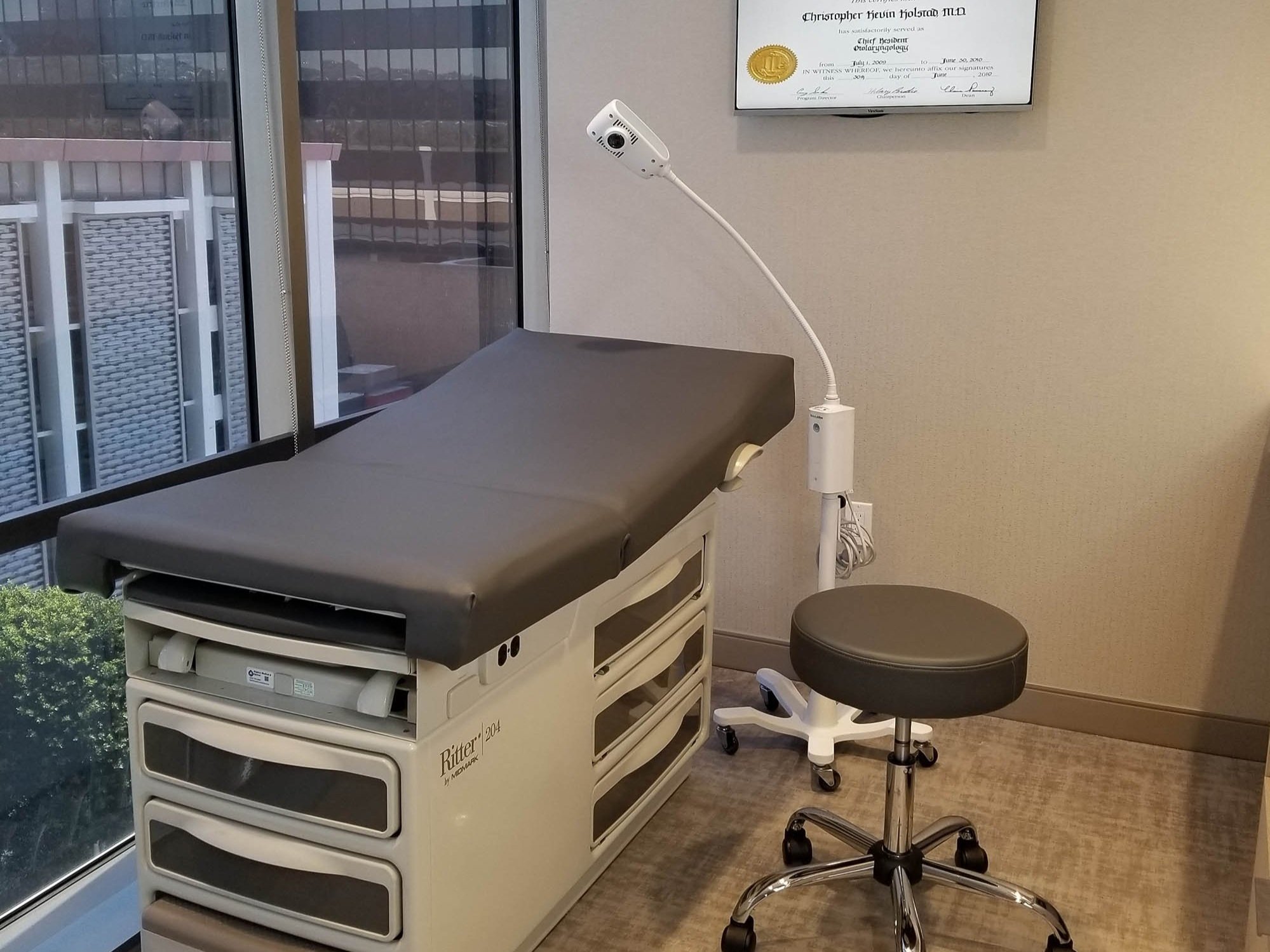 Medical examination room with exam table, stool, light, window view, and diploma on the wall.