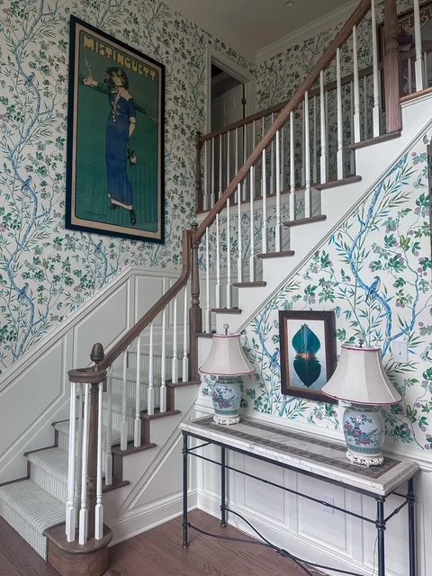 This elegant stair hall showcases luxury residential interior design that blends classic architecture with curated, high-end finishes. A traditional staircase with warm wood treads and crisp white balusters is elevated by hand-painted botanical wallp