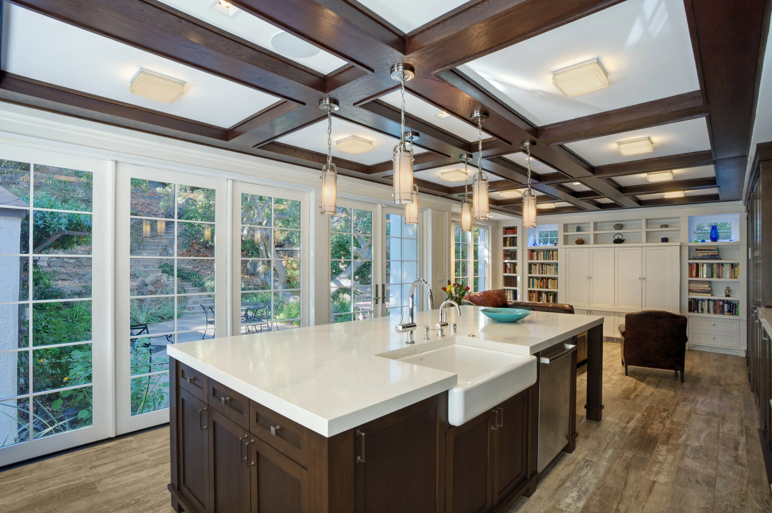Bright kitchen with large white countertop at the island, dark wood cabinets, and built-in bookshelf with books. Large windows and glass doors overlooking a garden with outdoor seating. Ceiling has wooden beams and suspended lights at the island.