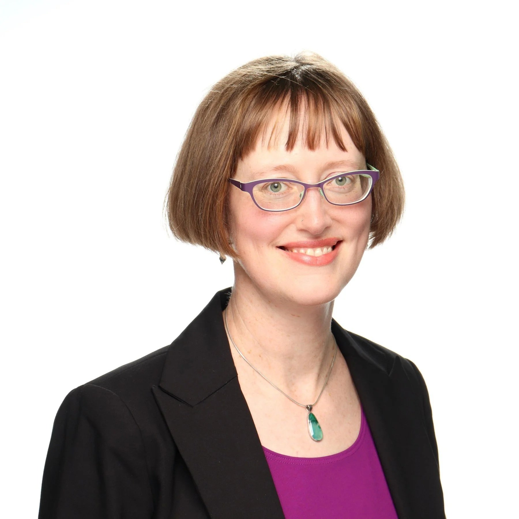 A woman with short brown hair, glasses, wearing a purple top, black blazer, and a green pendant necklace, smiling at the camera against a white background.