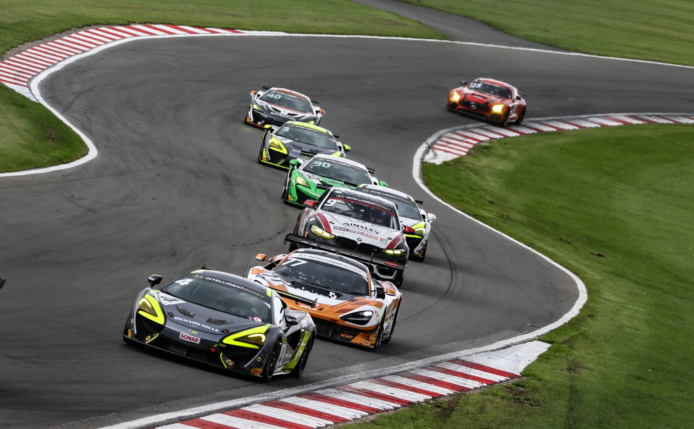 Race cars rounding a track corner during a competitive motorsport event, with green grass on the sides.