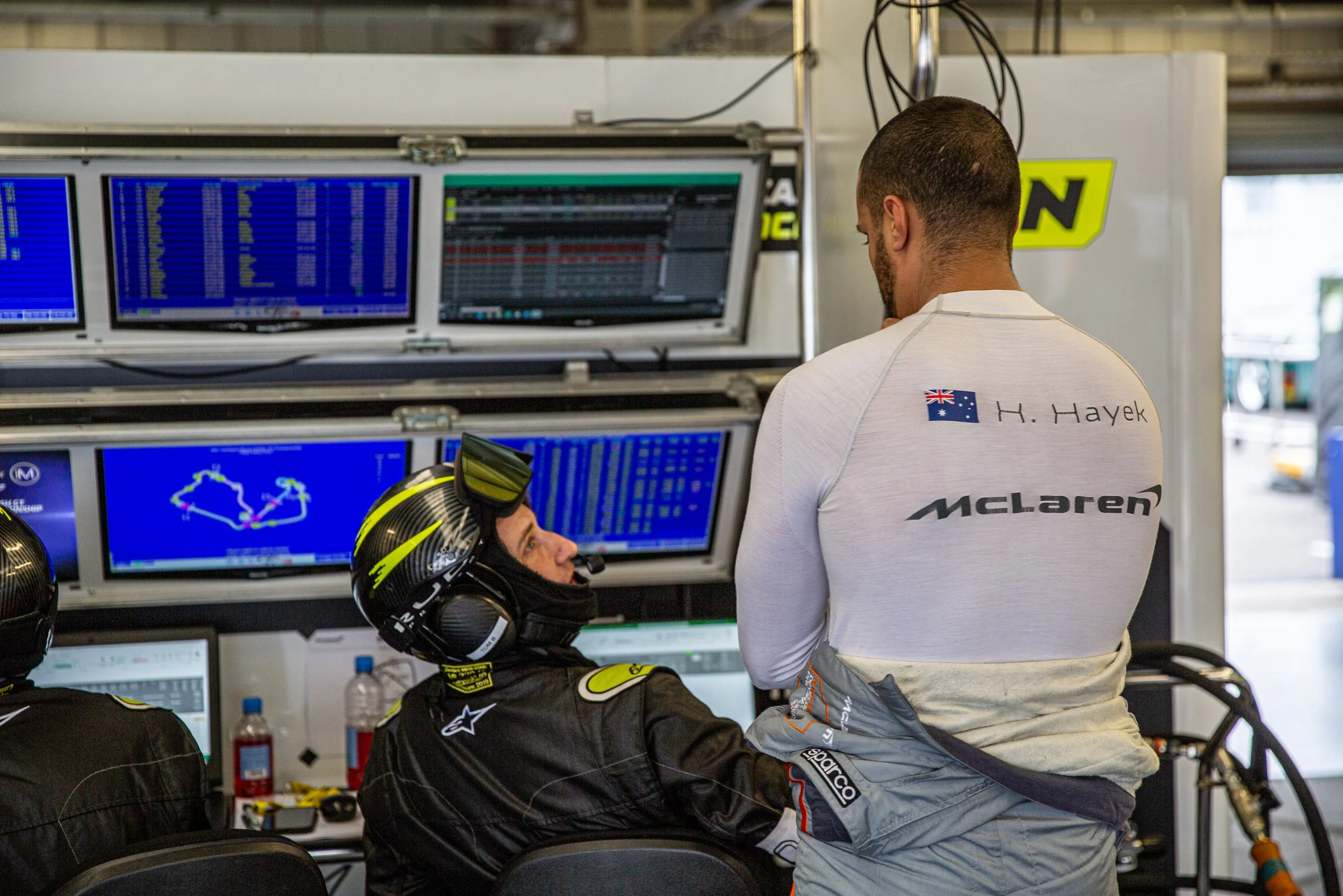 Race car driver in McLaren shirt talking to team members in pit garage with monitoring screens.