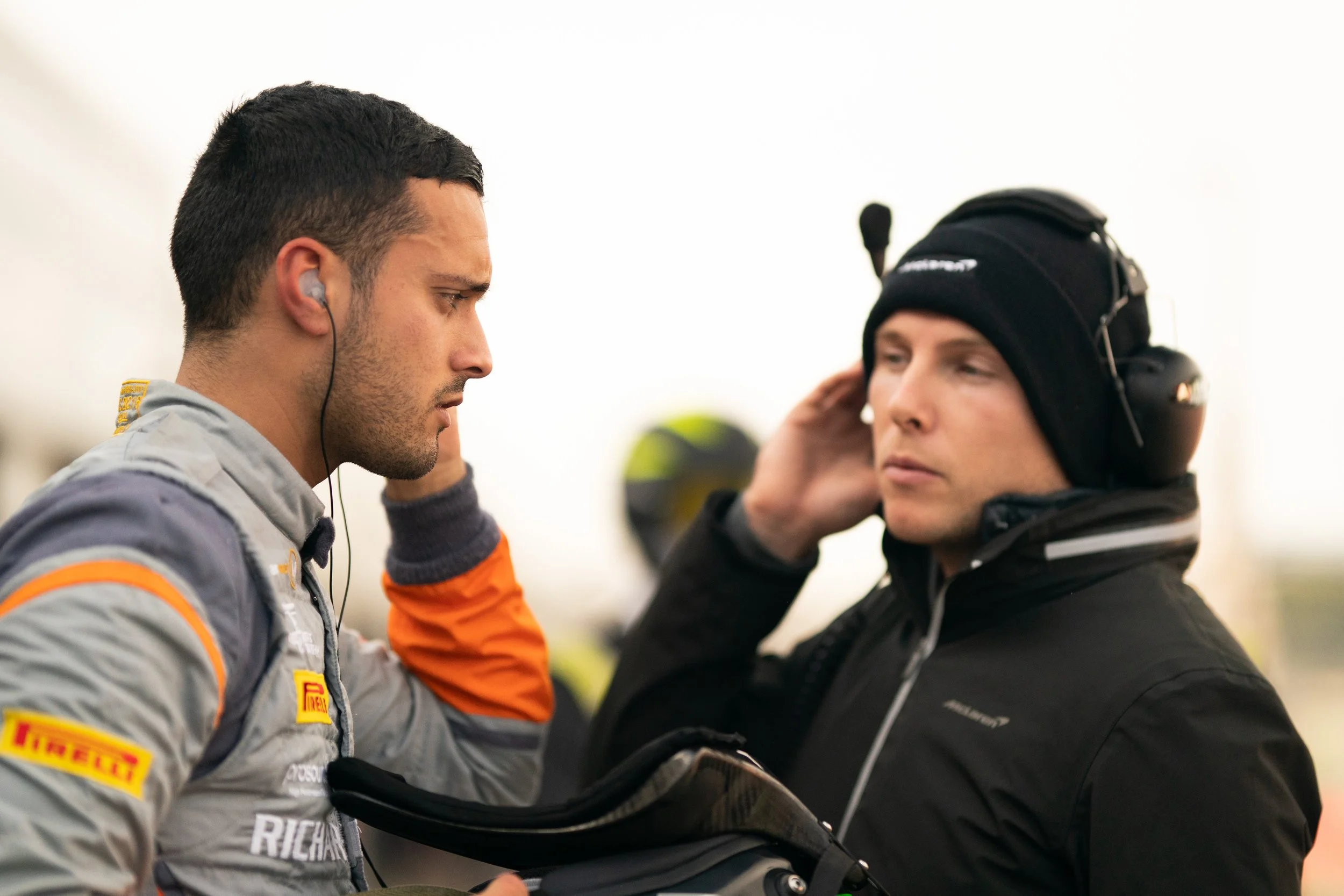 Racing driver in Pirelli-branded suit and team member with headset at racetrack.