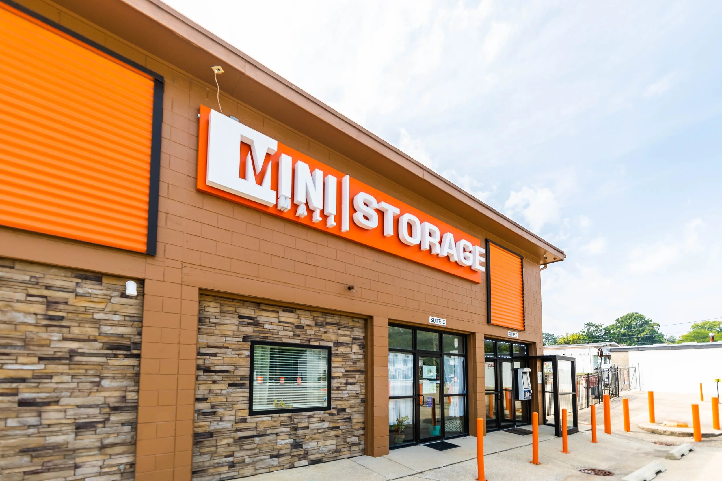 Exterior of a storage facility with a large orange sign that reads 'Mini Storage' on a brick building with orange accents and a stone lower wall, under a partly cloudy sky.