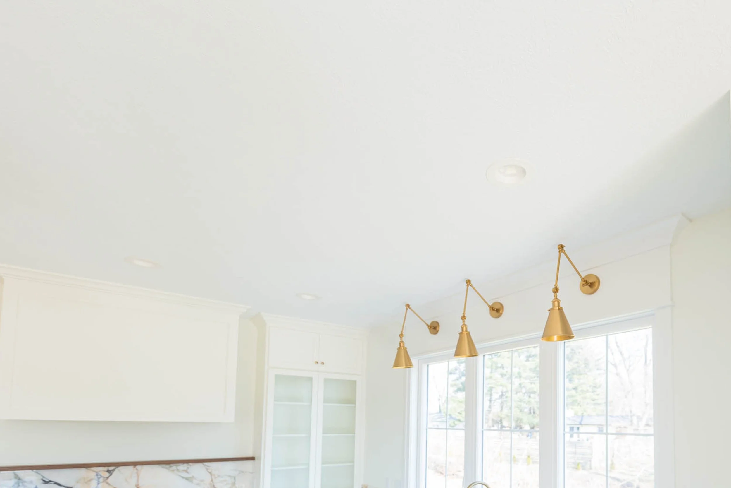 Bright kitchen interior with white walls and ceiling, featuring three gold pendant lights, large windows, and white cabinetry.