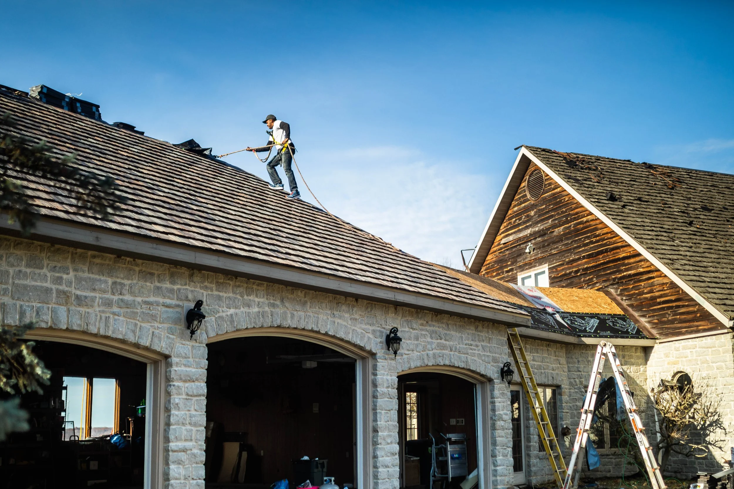 A person working on roof shingles of a house, using safety harness and rope, with ladders leaning against the house during daylight.