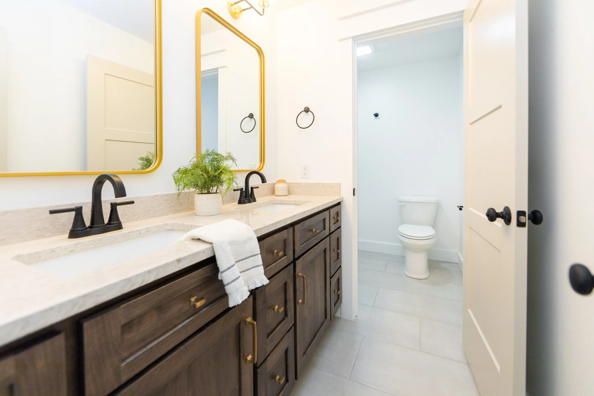 Bathroom with double vanity sink with dark wood cabinets, beige countertop, two black faucets, a potted plant, and mirrors with yellow frames. Visible toilet behind the open door, white walls, and tile floor.