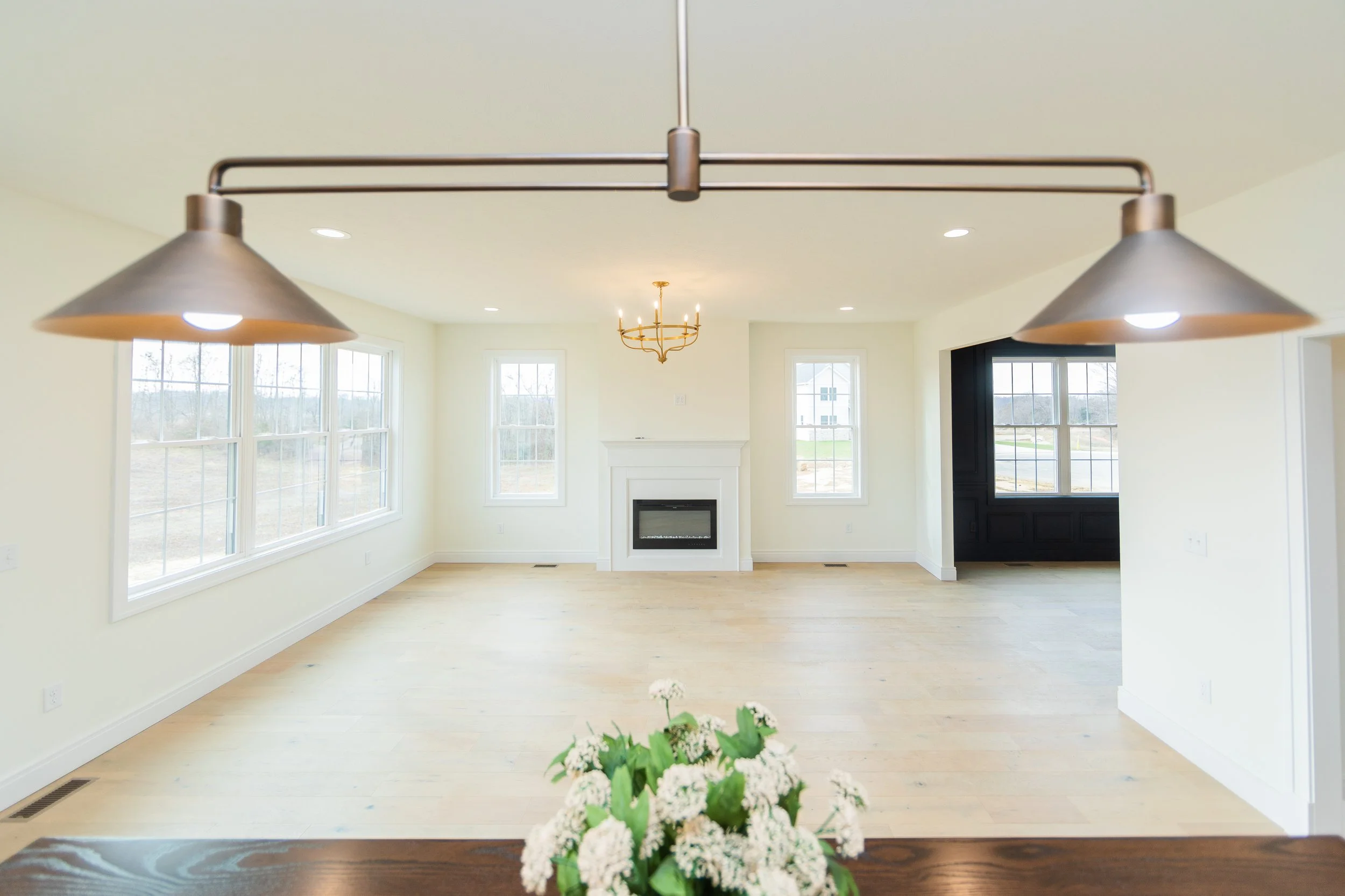 Empty living room with large windows, a fireplace, hardwood floors, and a chandelier, viewed from a countertop with a flower arrangement.