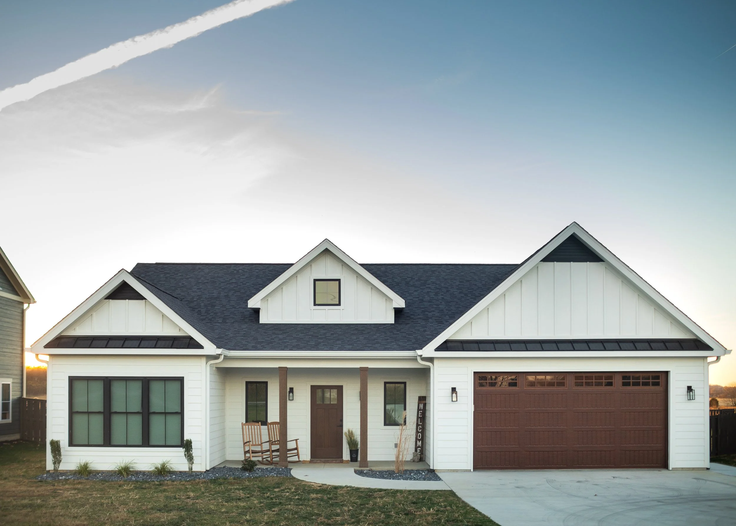 Modern white house with black roof and brown garage door, front porch with two wooden rocking chairs, small plants and welcome sign, sunny evening sky.