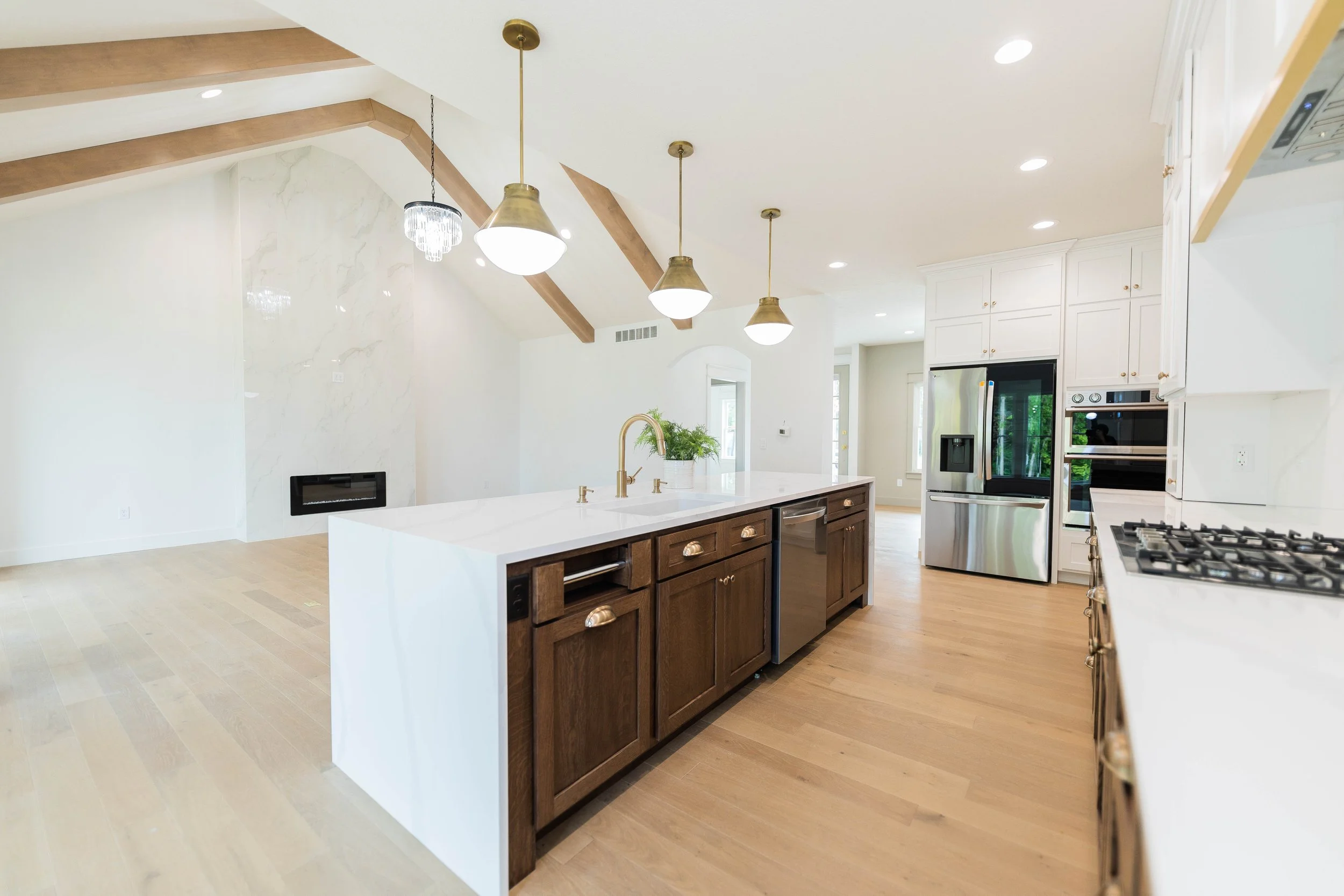 Modern kitchen with white countertops, wooden cabinets, stainless steel appliances, and a spacious island with a gold sink, under a ceiling with wooden beams and pendant lighting.