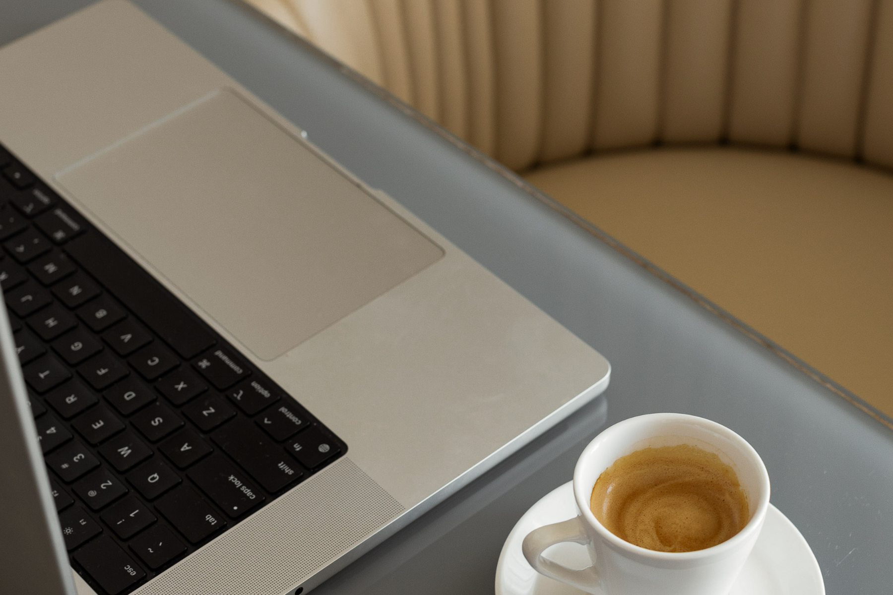 A cup of coffee and a laptop sit on a desk, a neutral colored chair pulled out.