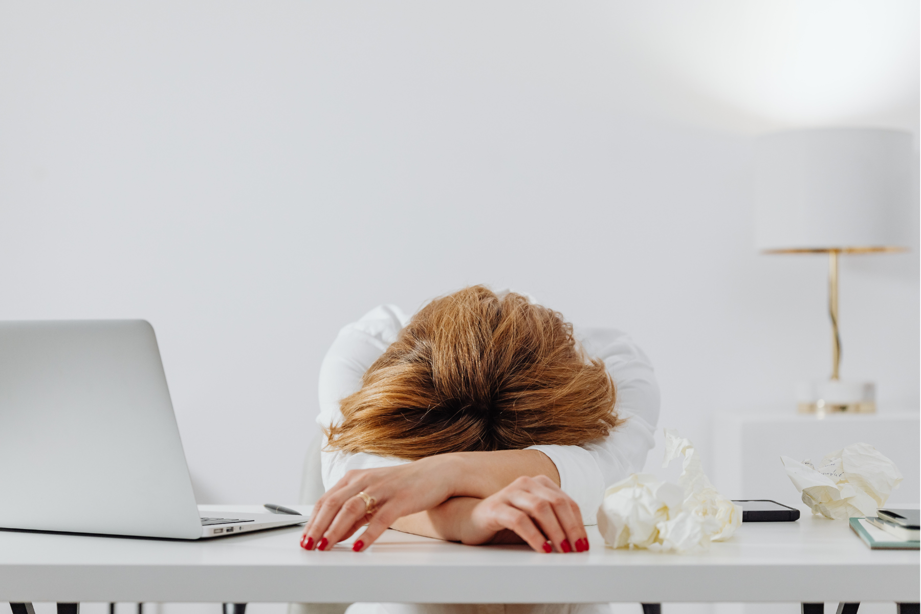 A woman is slumped face-first on her white desk, her head facing the camera and arms crossed on the tabletop