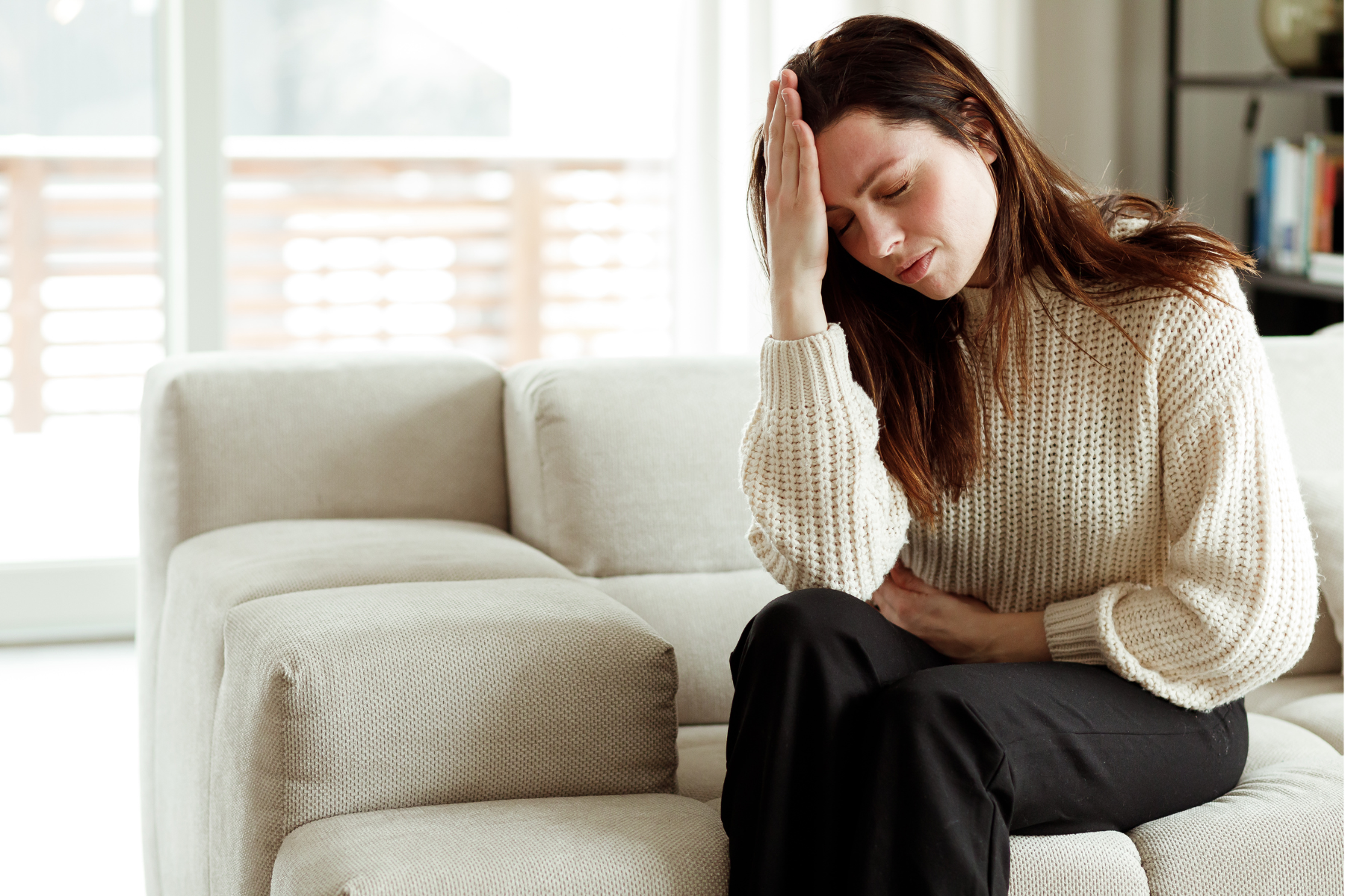 A woman sits on a couch, her hand to her head and her other hand on her stomach, her eyes closed.