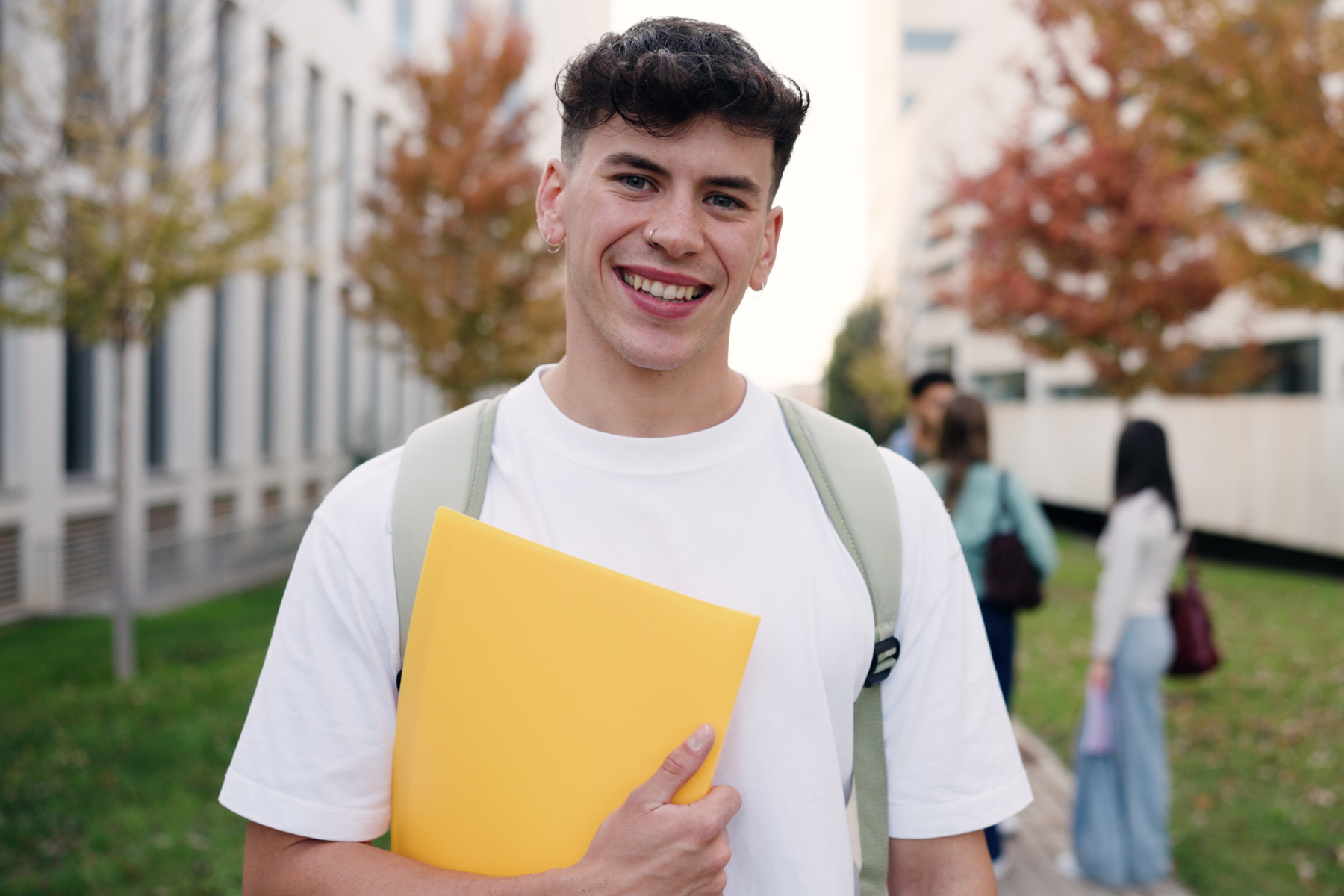 A college student stands on an outdoor path, framed by autumn trees. He is smiling and wearing a white shirt and sage green backpack, and carrying a yellow folder.