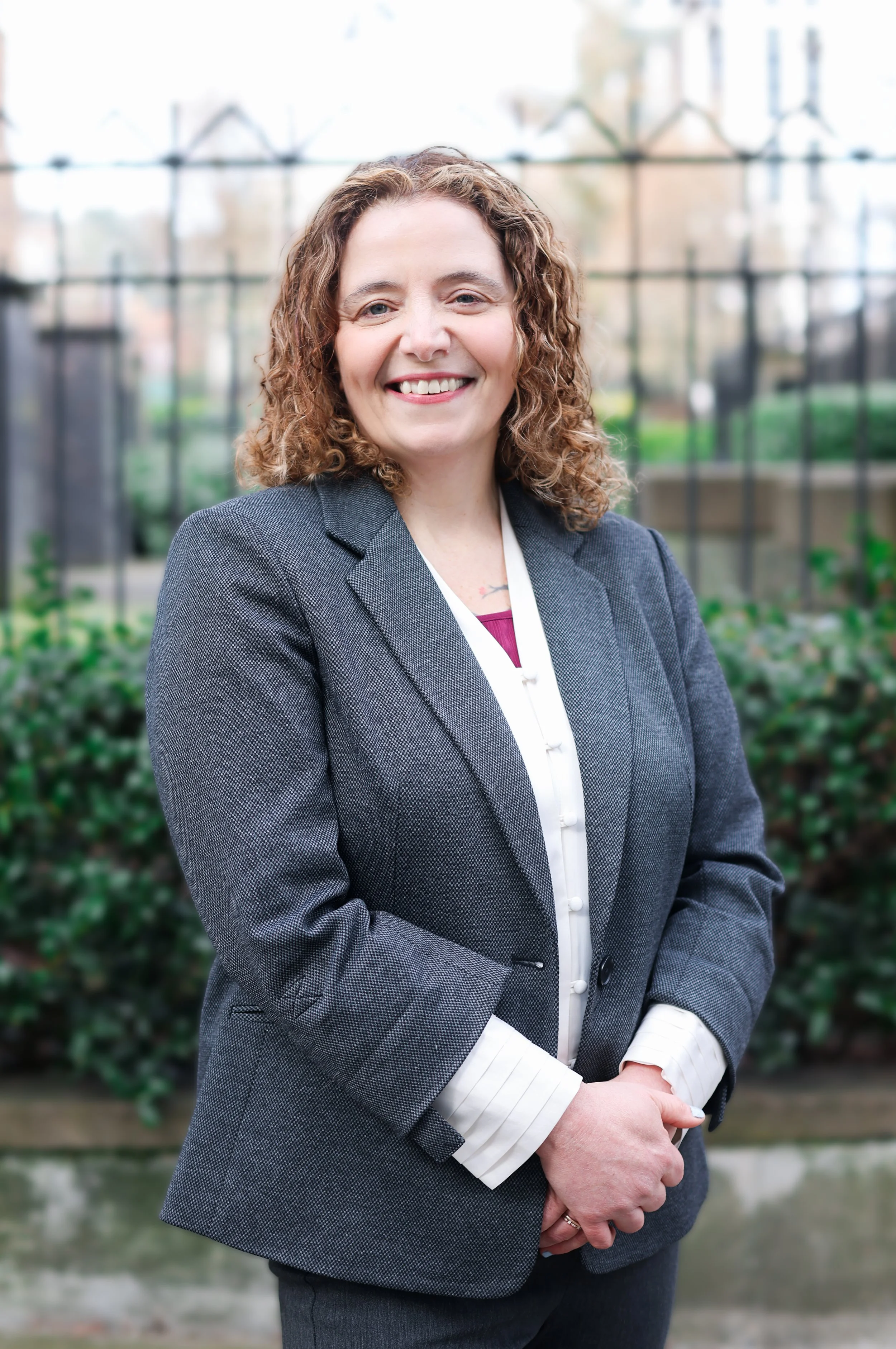 A photo of Alesin Sinclair standing outside in front of a gate. She has short, curly hair and is smiling, wearing a blazer.