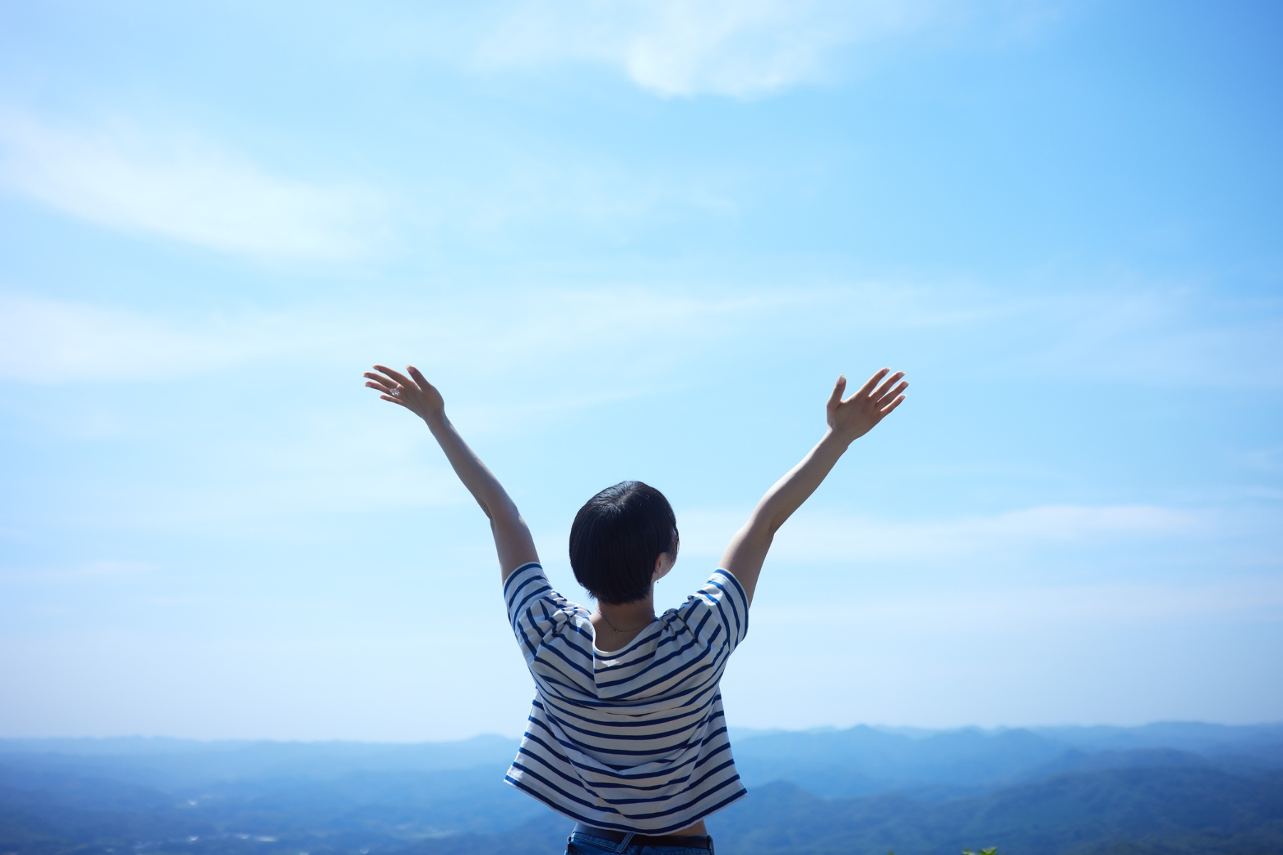 A woman photographed from behind, raising her hands to the sky as she overlooks a mountain range.