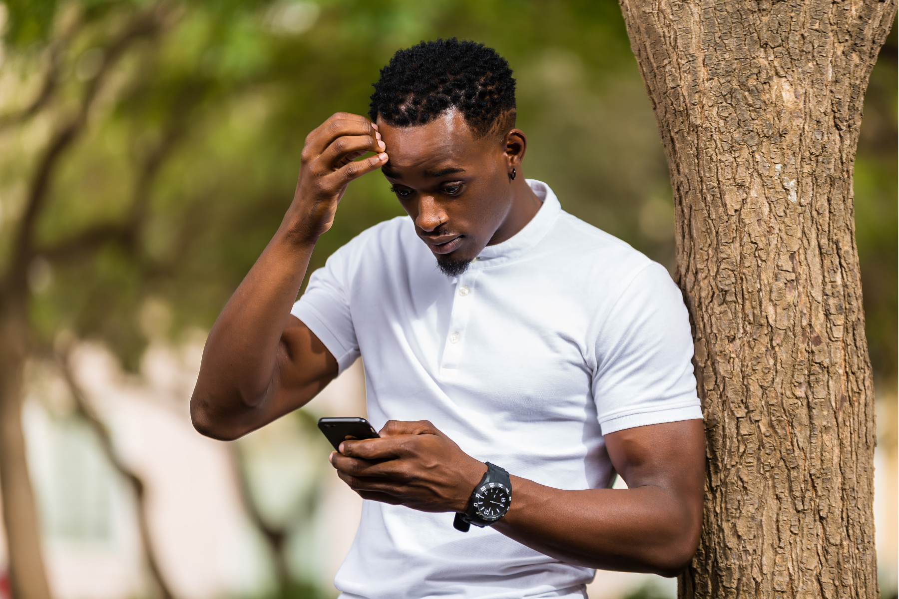 A man wearing a white shirt leans against a tree, looking down at his phone, his expression stressed