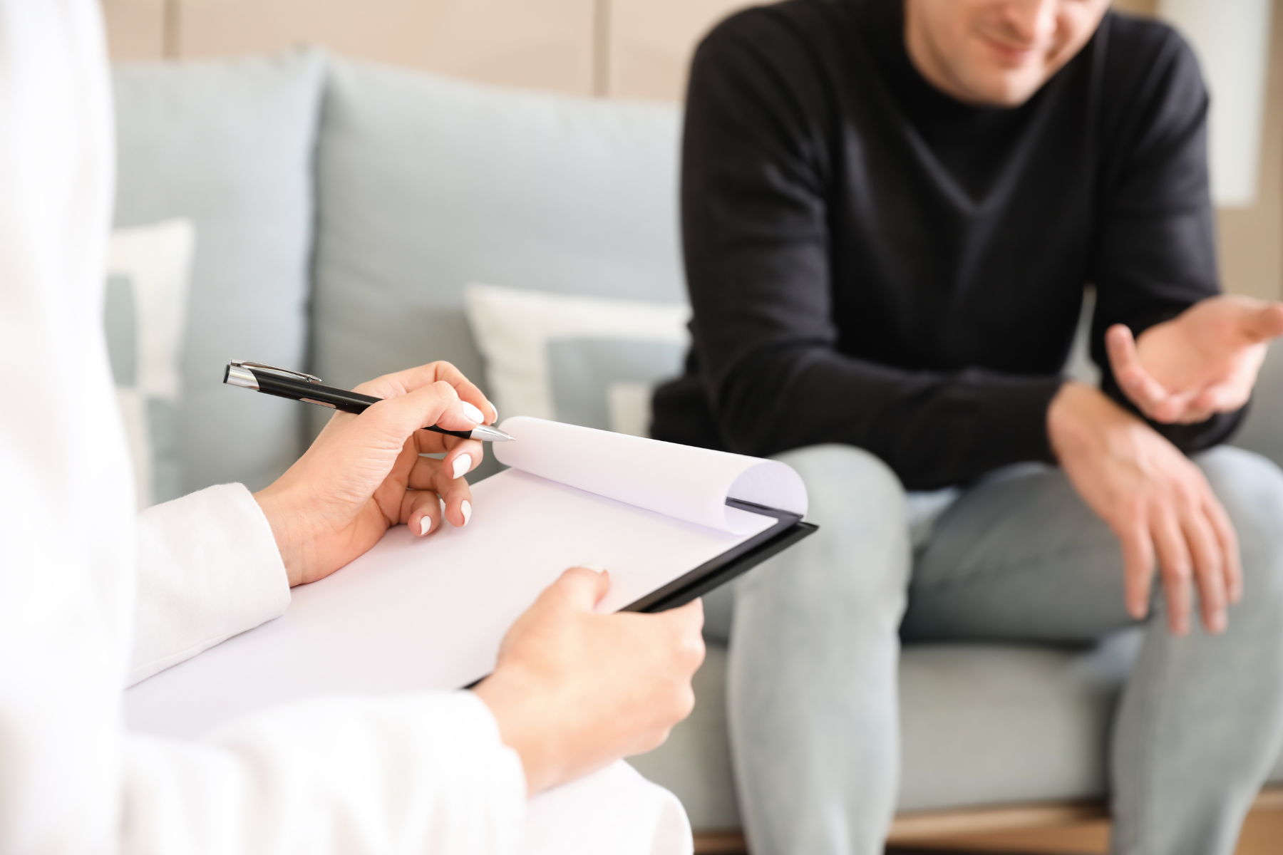 A man sits on a couch, speaking with a therapist, who is taking notes on a clipboard.