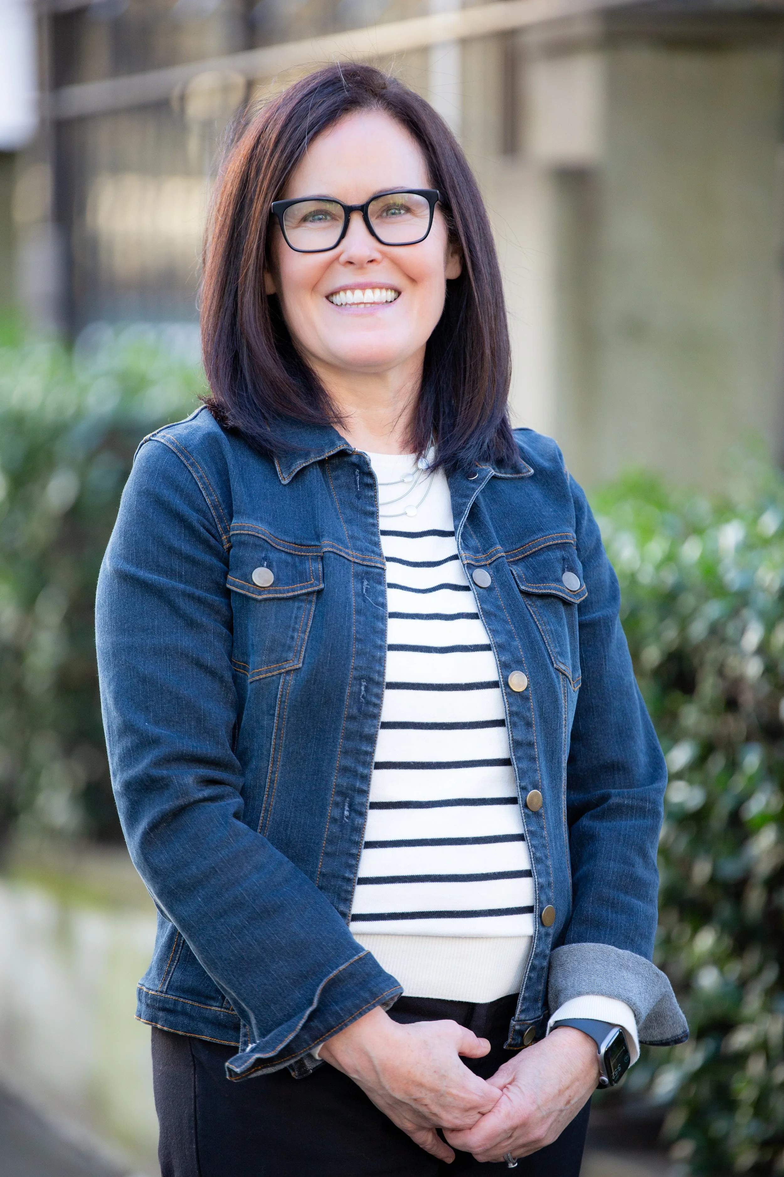 Jennifer Clark, a Caucasian woman with dark hair, stands, smiling. She is wearing a black and white striped shirt and a denim jacket.