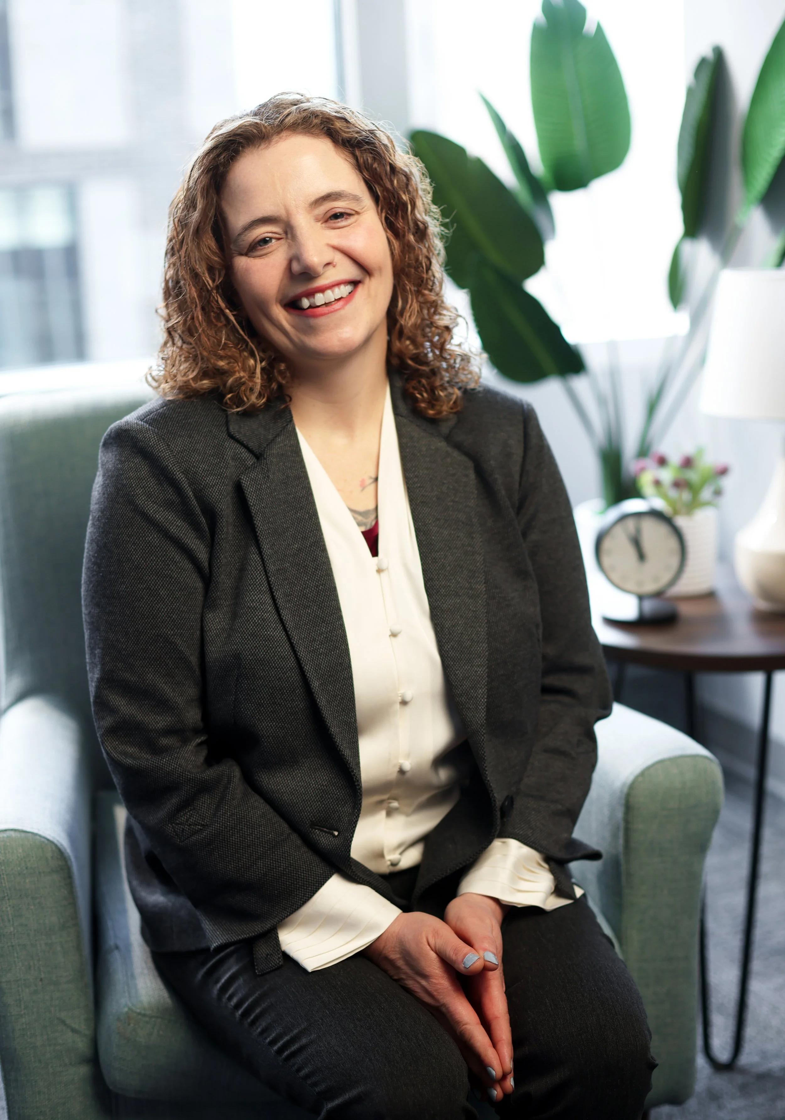 Alesin Sinclair sits in an armchair, smiling. She has short, curly hair and is wearing a blazer.