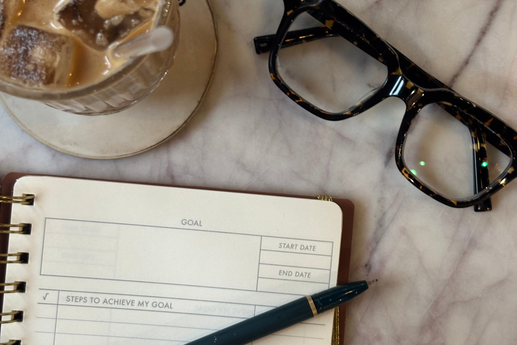 A pair of glasses, a cup of coffee, and a goal-setting notebook sit on a marble tabletop.