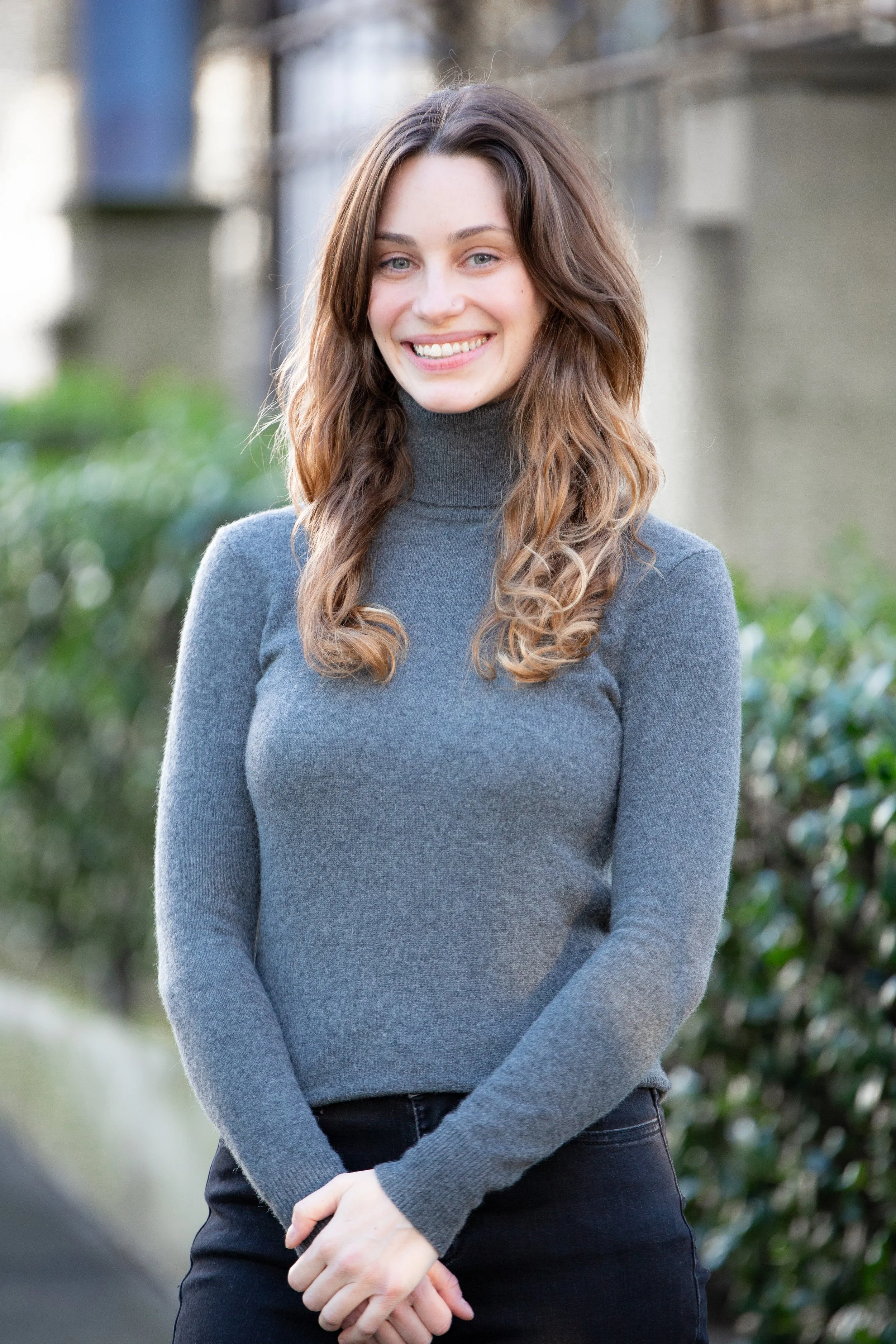 Leah Marinakis, a Caucasian woman with brown curly hair, smiles. She is wearing dark pants and a gray turtleneck sweater.