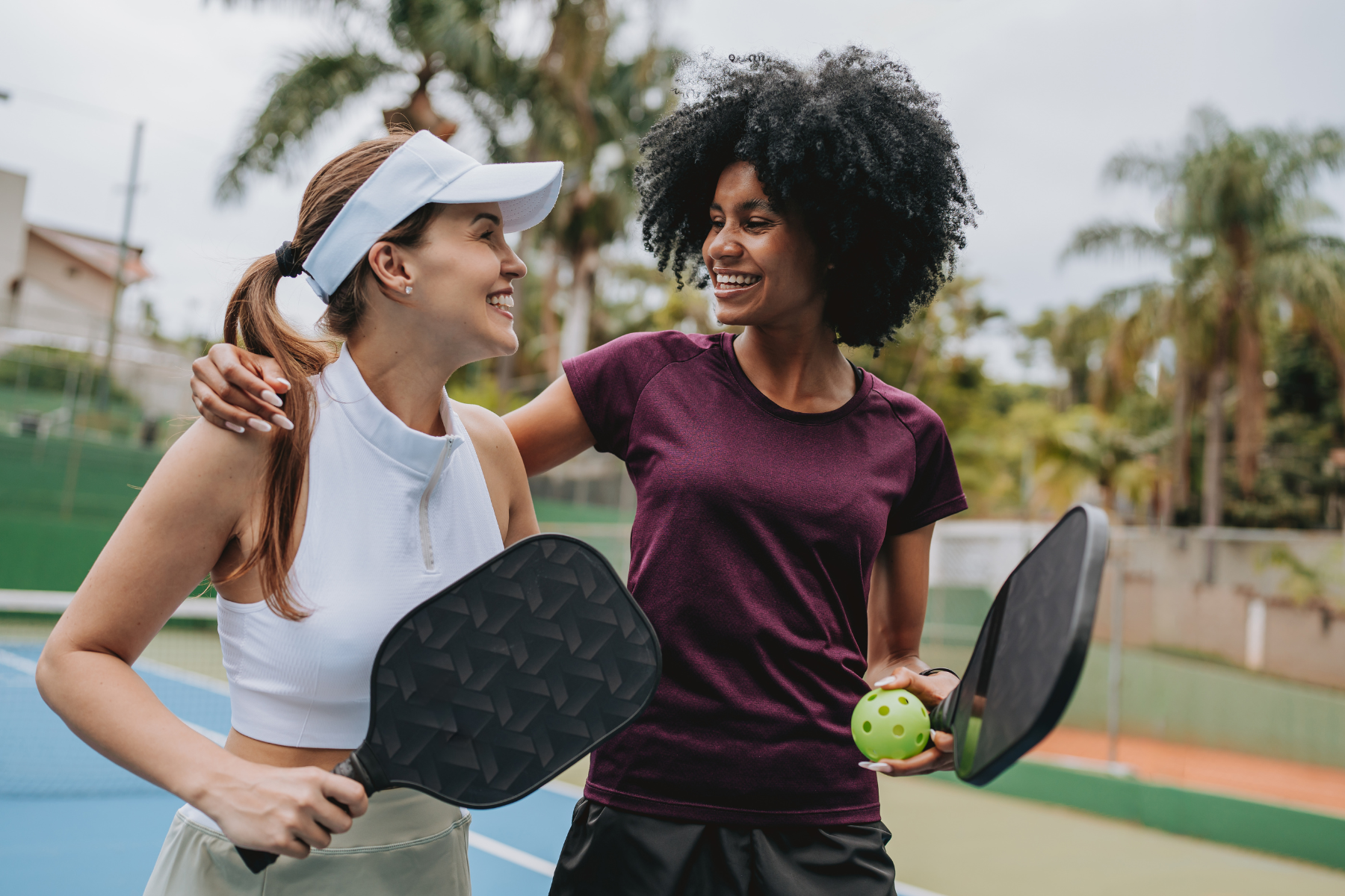 Two women are on the pickleball court together. One woman has an arm around the other woman's shoulders.