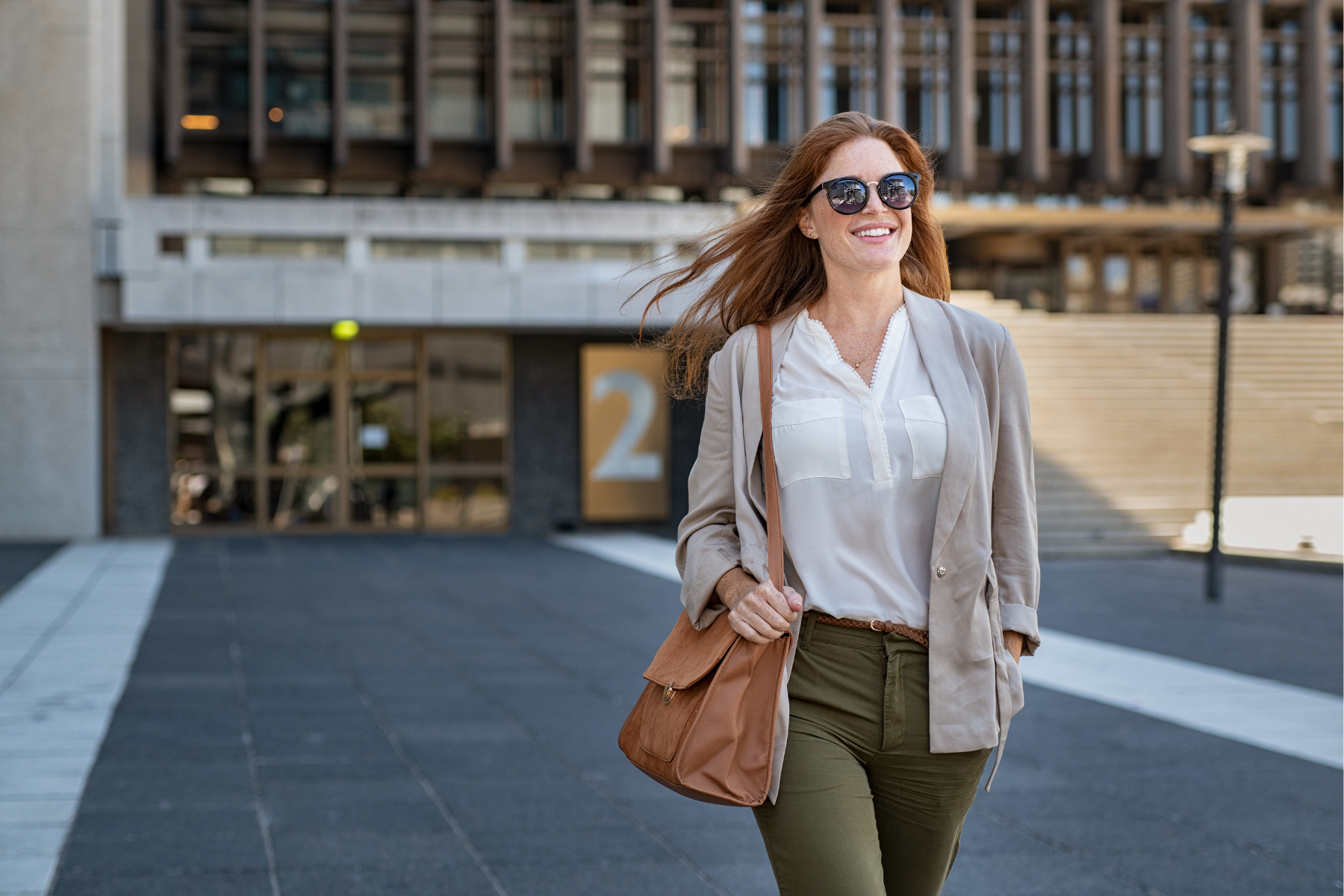 A redheaded woman walks confidently outside, her hand on her shoulder bag.