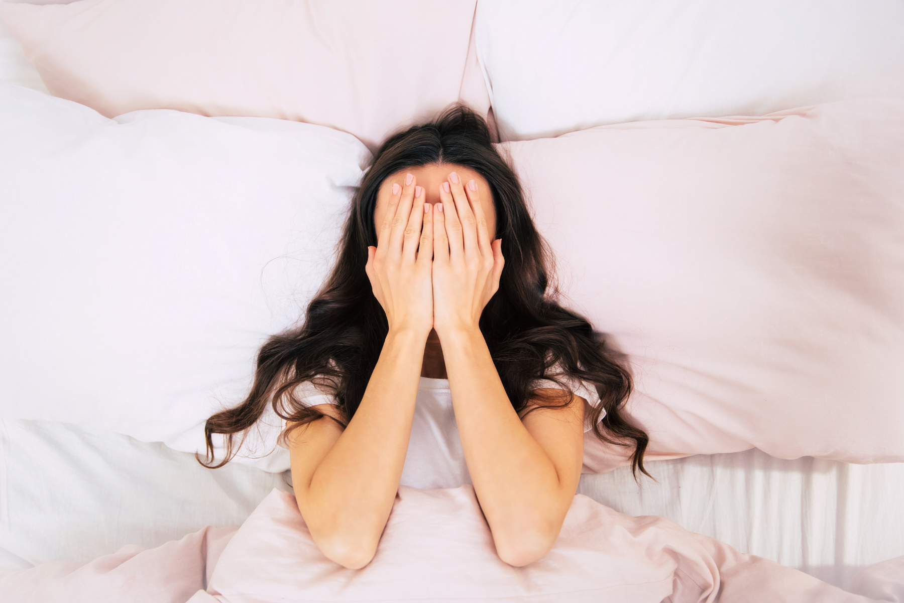 A woman with dark, curly hair lays in bed on a pink pillow with her hands covering her face.