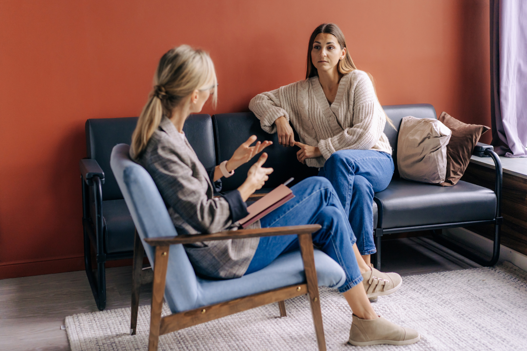 A woman sits on a couch, listening to a therapist in a nearby chair.