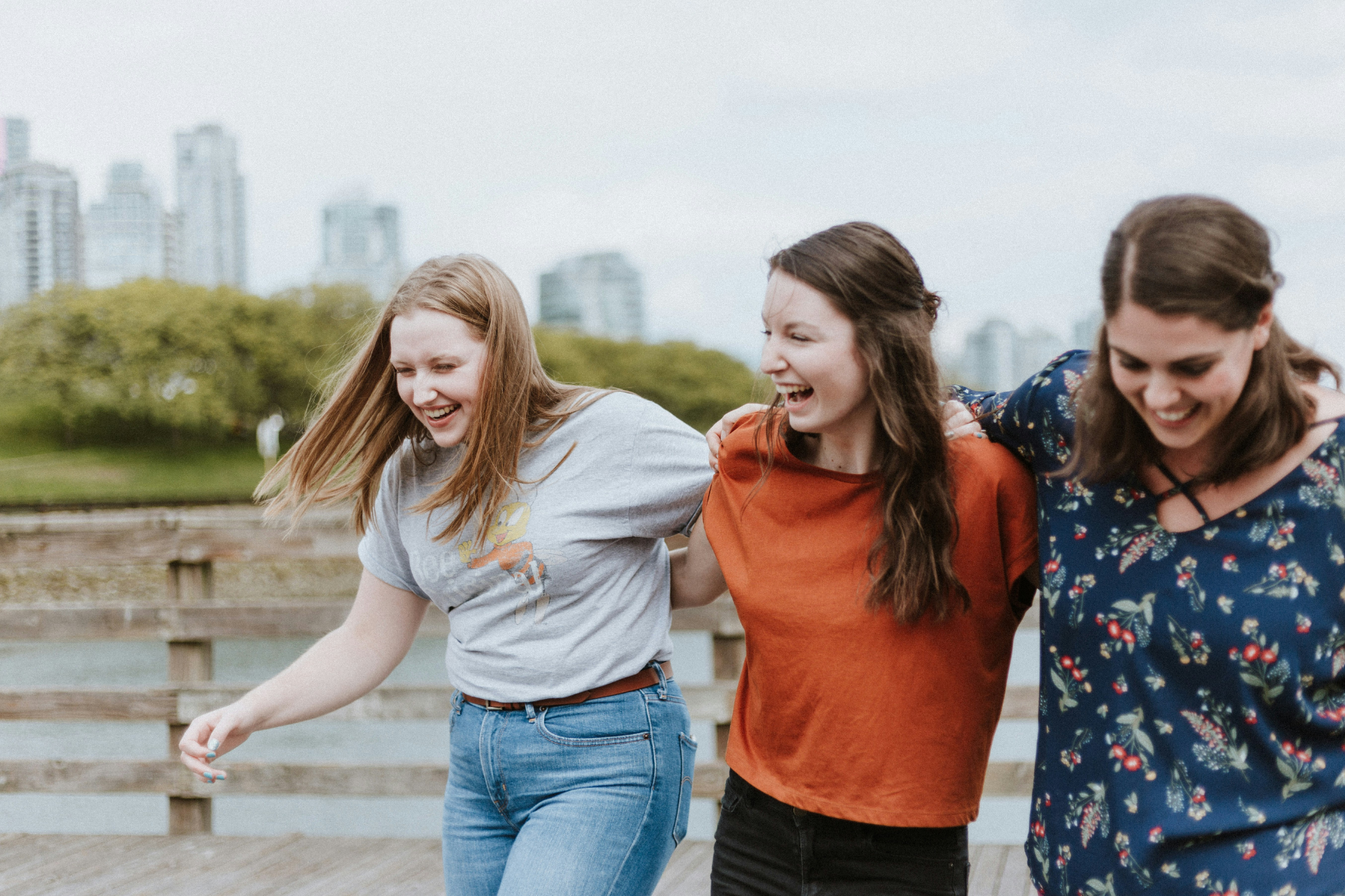 Three women walk along a city park, arms around each other, laughing