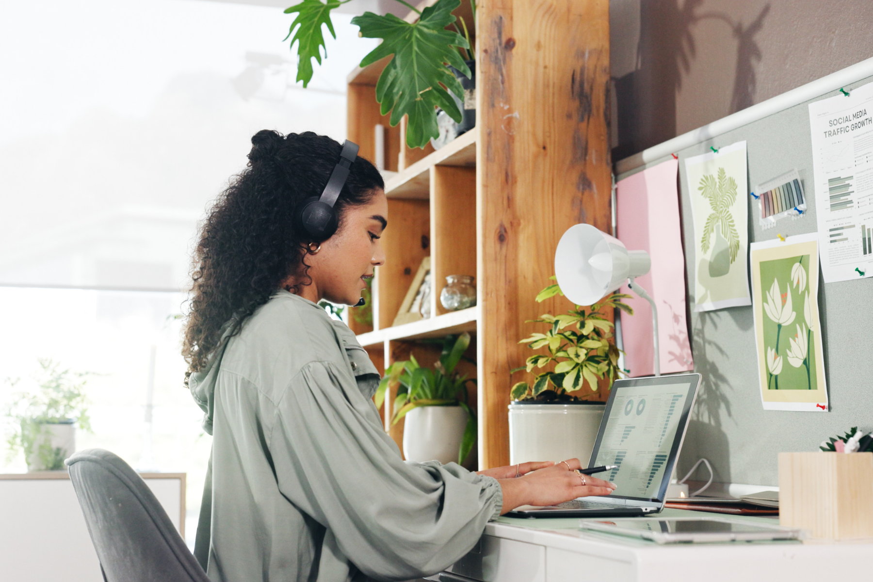 A woman with dark curly hair sits at her laptop, working with headphones on.