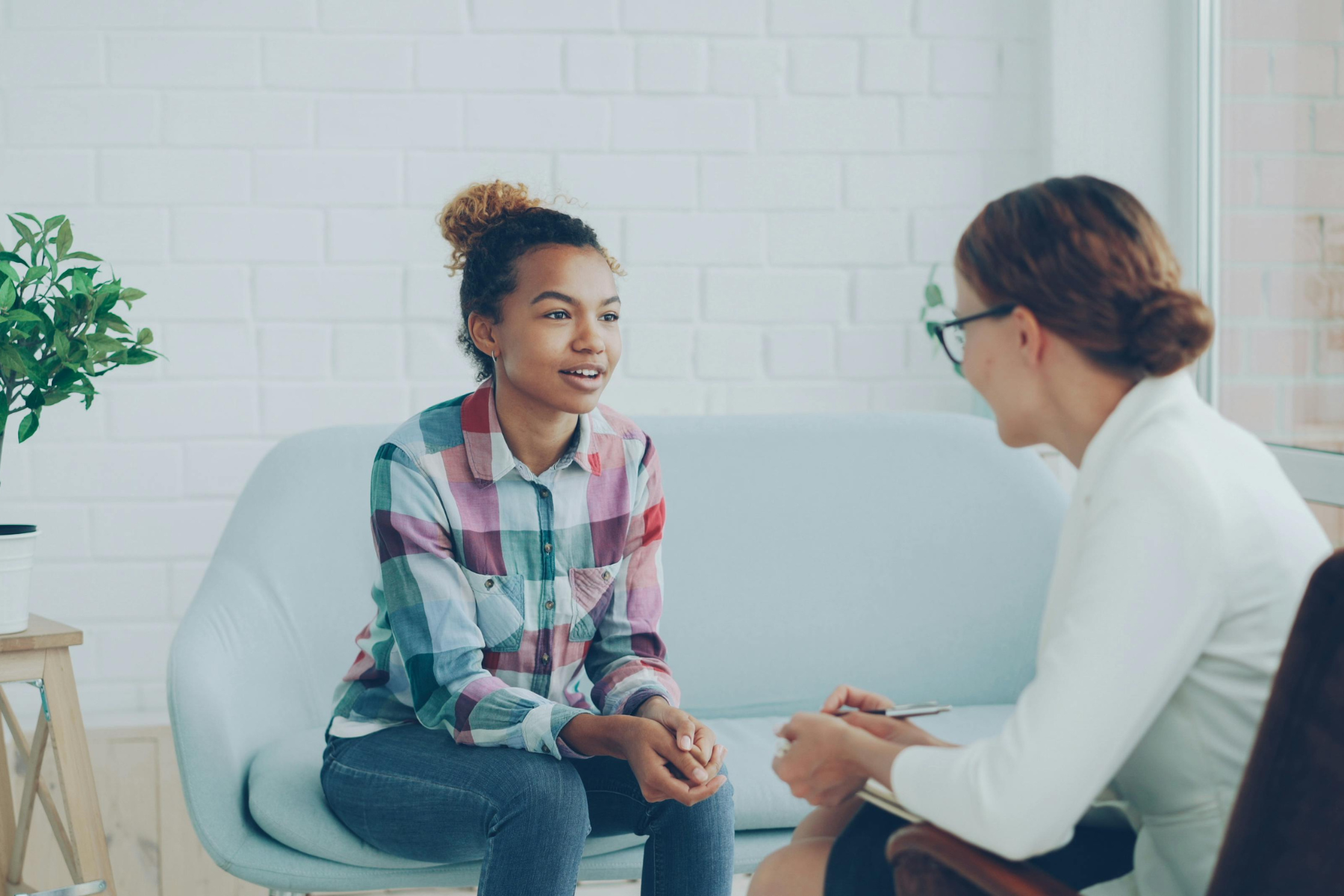 A young woman wearing a colorful flannel sits on a blue couch, talking with her therapist.