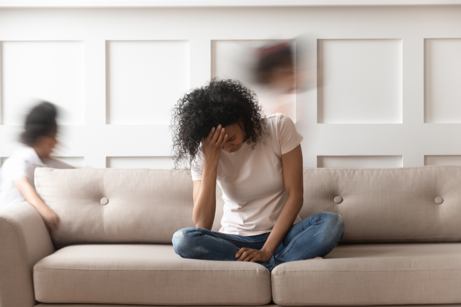 a woman sits on the couch, overwhelmed and burying her head in her hands while life continues on in a blur around her