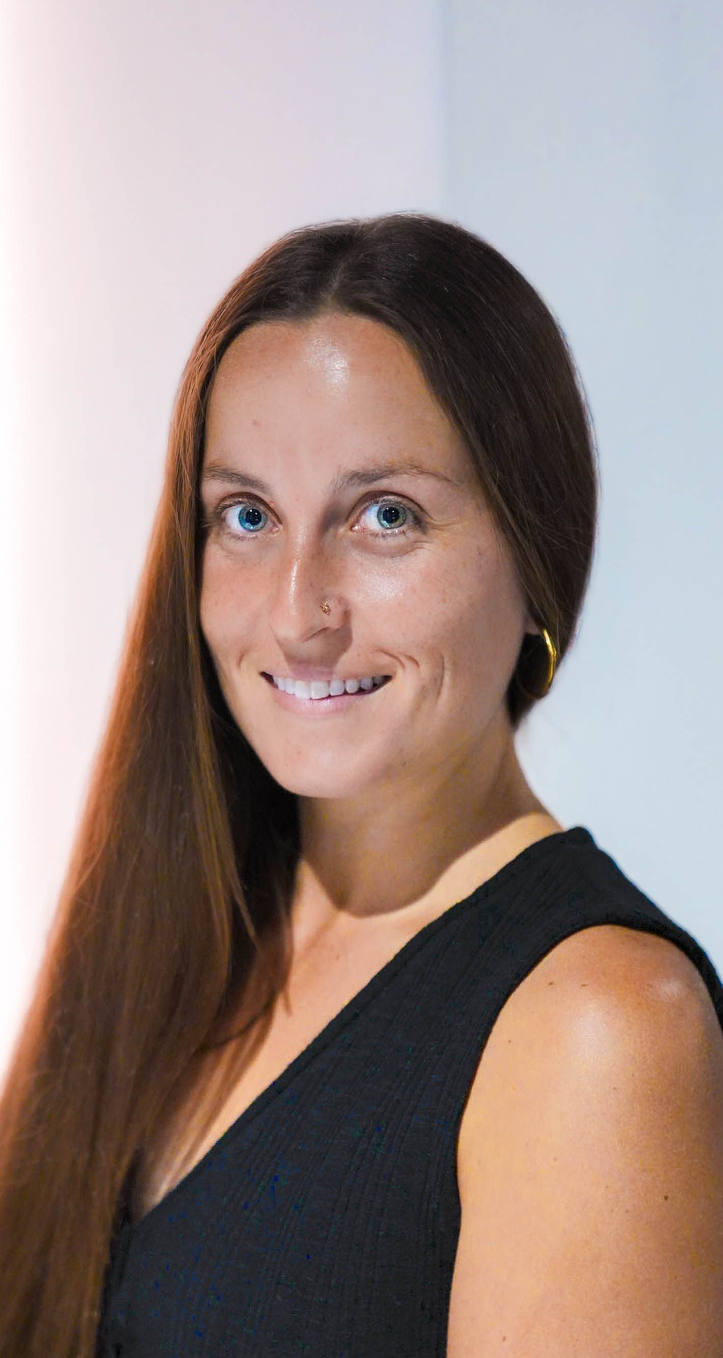 Close-up portrait of a woman with long brunette hair, blue eyes, a nose ring, and gold hoop earrings, wearing a black sleeveless top, smiling.