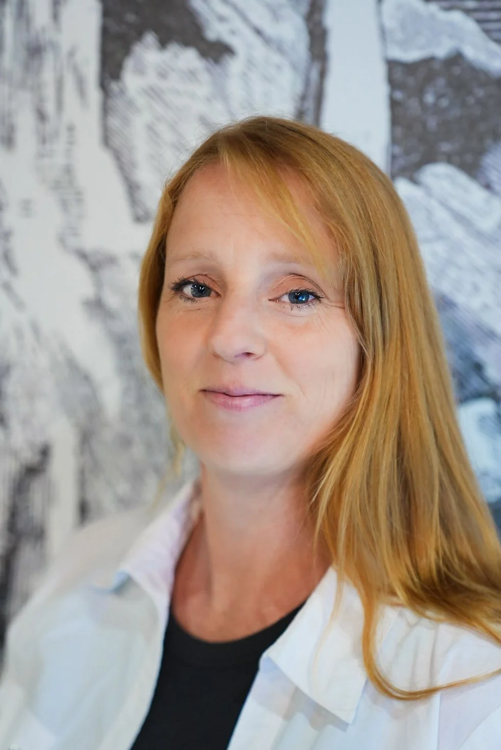 Close-up portrait of a the owner of kind body wellness with long red hair, blue eyes, and fair skin, smiling slightly, wearing a white shirt over a black top, against a blurred background of black-and-white artwork.