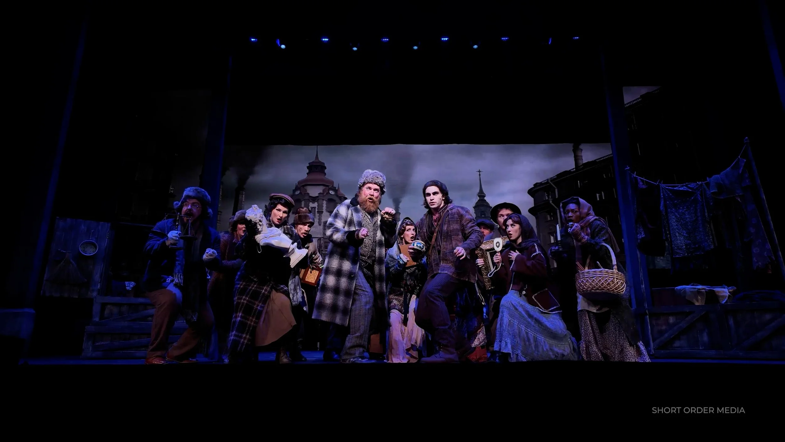Stage scene with actors dressed in vintage clothing, performing in front of a backdrop of city buildings with smoke. The actors appear to be engaged in a theatrical play, some holding props like phones and baskets.