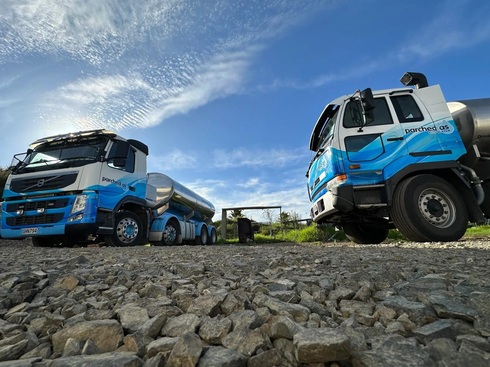 Two Parched As Water trucks with blue and white paint parked on gravel ground under a partly cloudy sky.