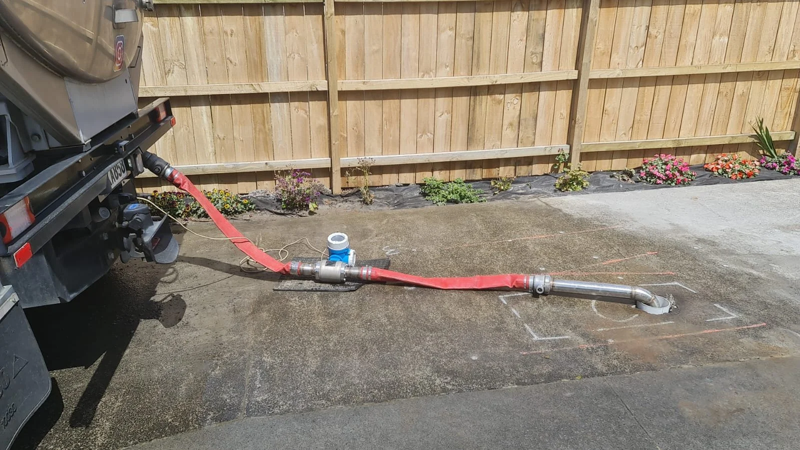 Parched As water truck red hose connected to a blue flow meter, on a concrete surface next to a wooden fence with small flowers plantations.