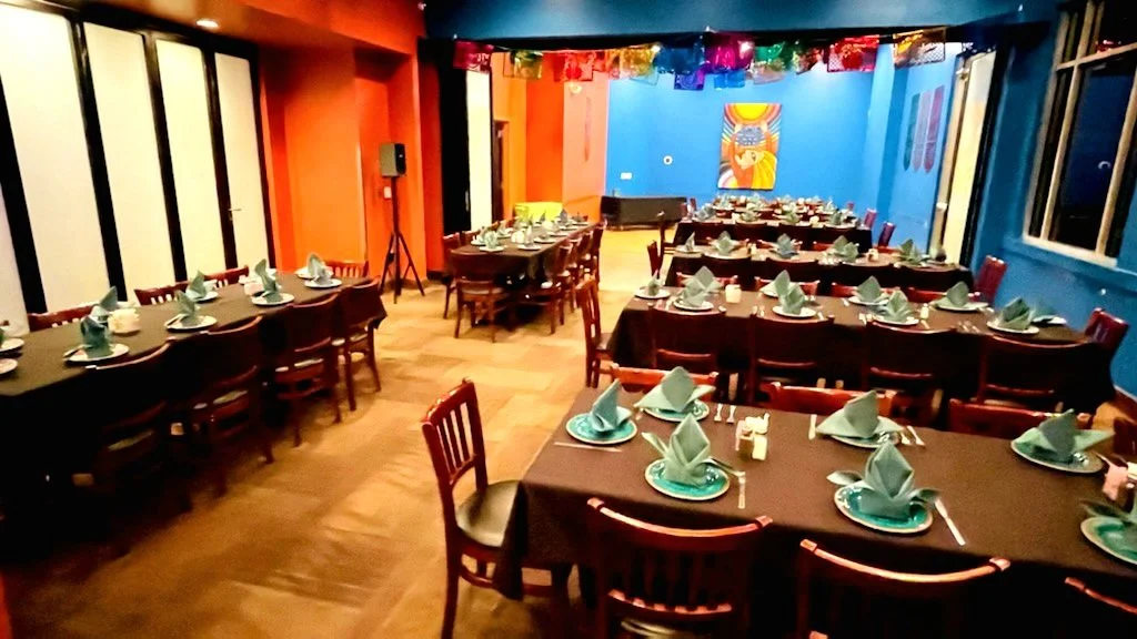 Indoor party or event space decorated with colorful bunting, with tables set with folded napkins, plates, and utensils, arranged for a meal.