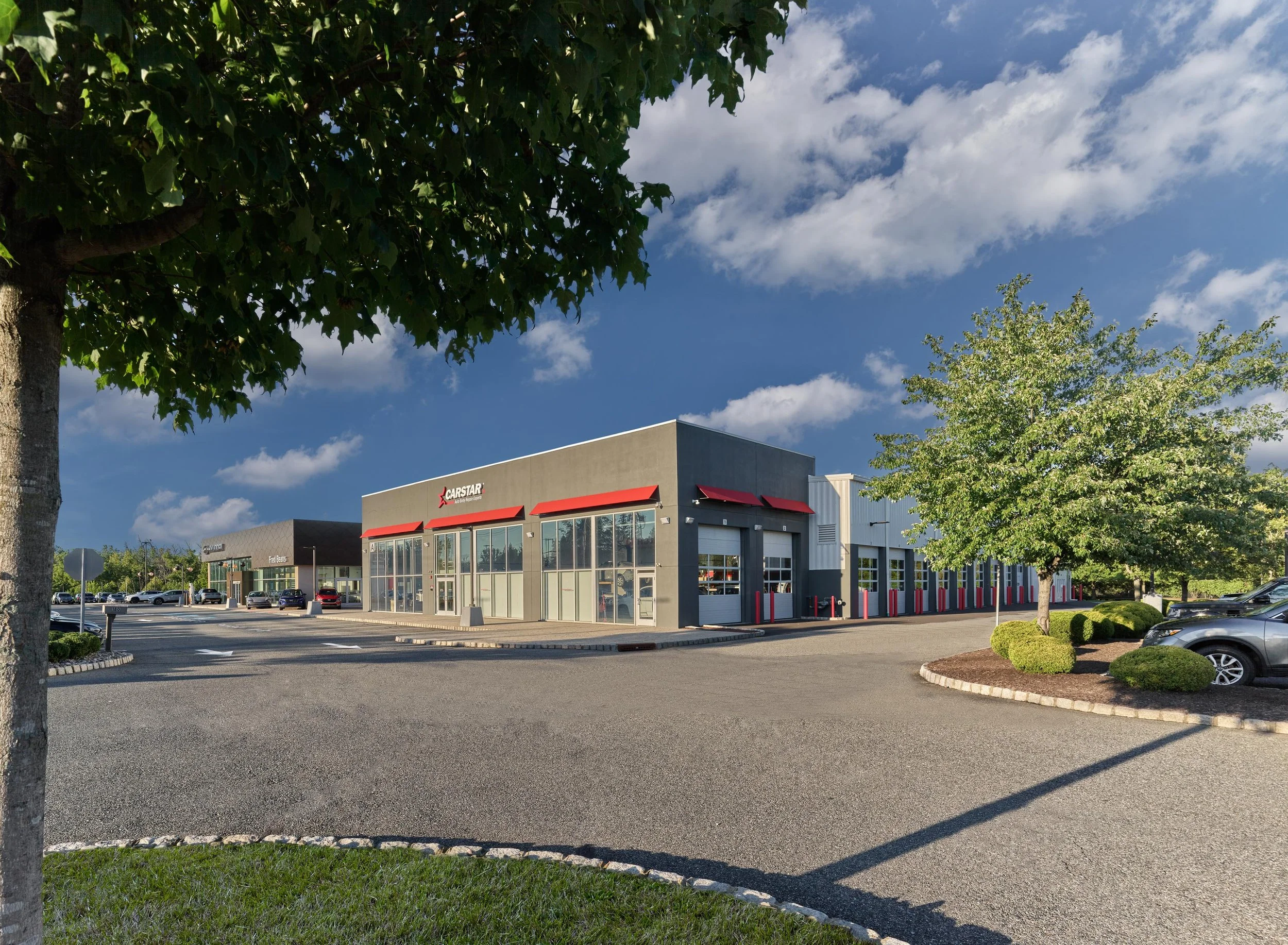 A car dealership building with multiple glass windows and a sign that reads 'Carstar' with a red logo. The parking lot is mostly empty with a few cars visible, and there are trees and a clear blue sky with some clouds overhead.