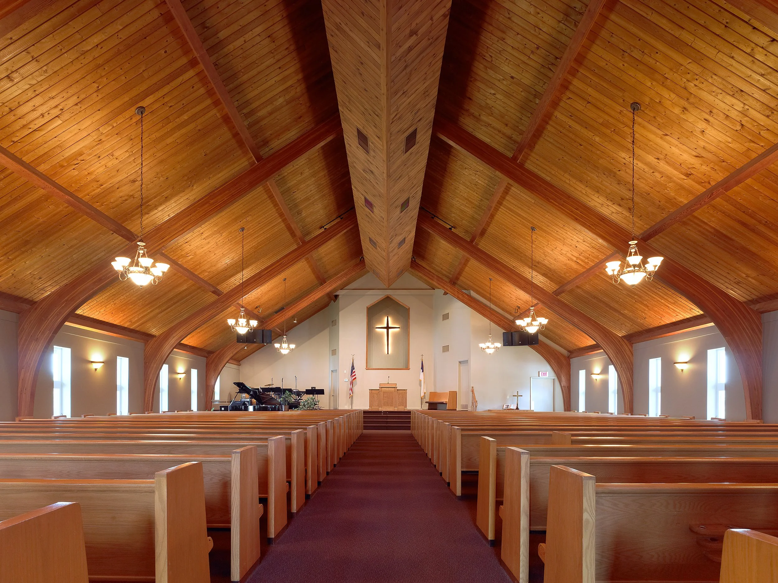 Interior of a church sanctuary with wooden pews, a raised altar with a cross, and a piano, under a high wooden ceiling with chandeliers hanging from it.