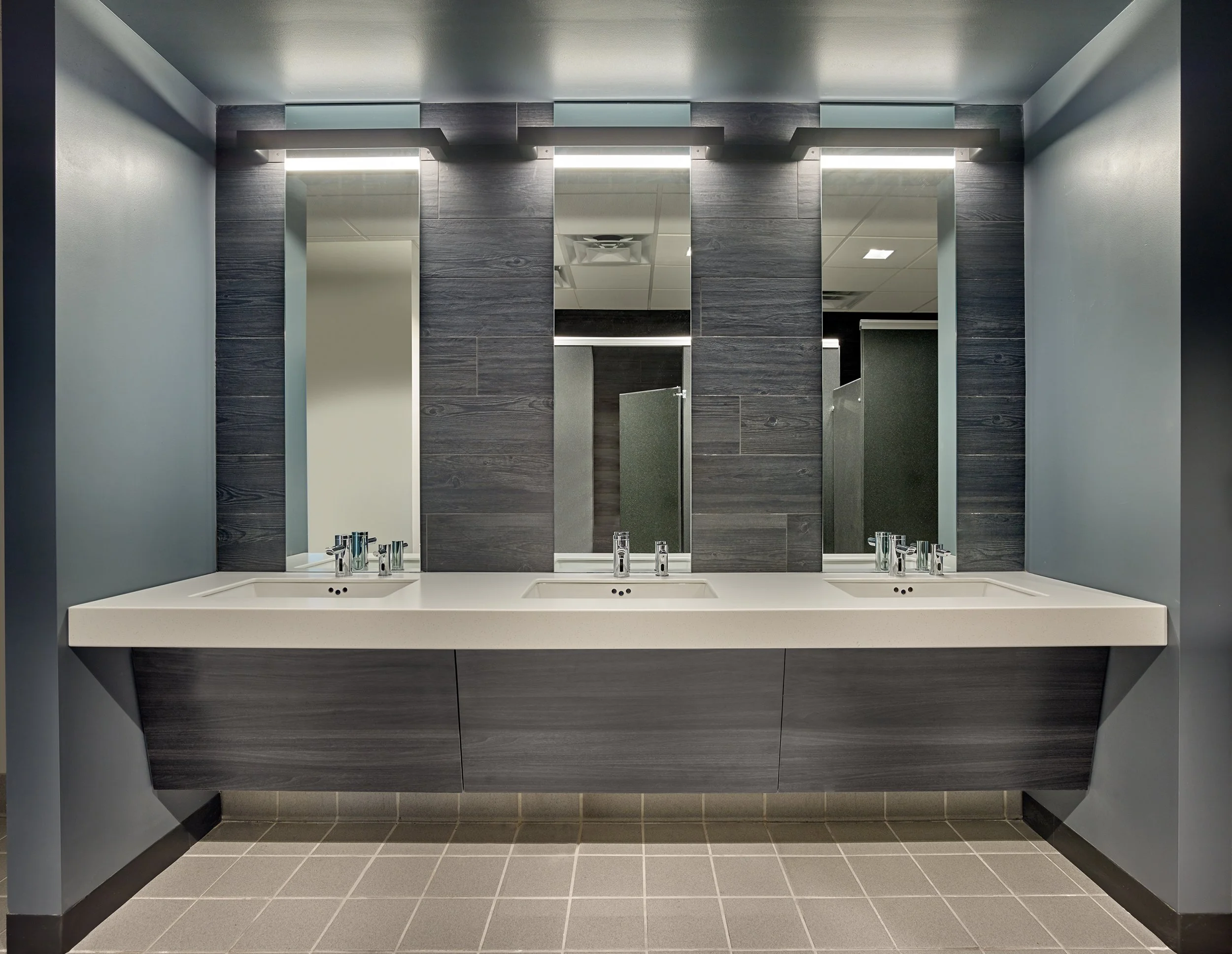 Public restroom sinks with three mirrors above them, dark wood paneling, gray tiled floors, and soap dispensers.