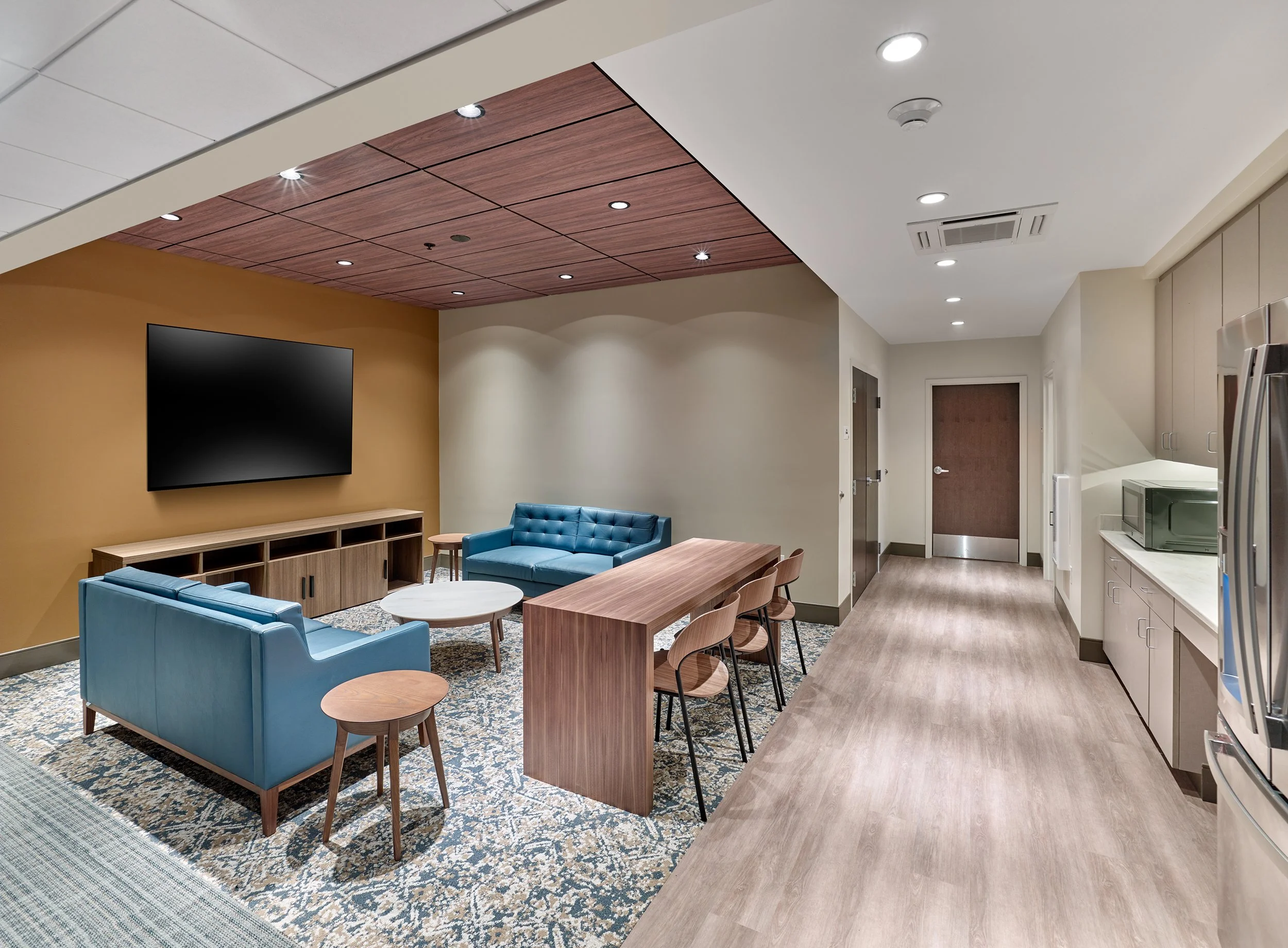 Modern dormitory lounge area featuring blue sofas, a wooden coffee table, and a vanity with a TV on the wall, adjacent to a kitchenette with microwave and refrigerator in a well-lit room.