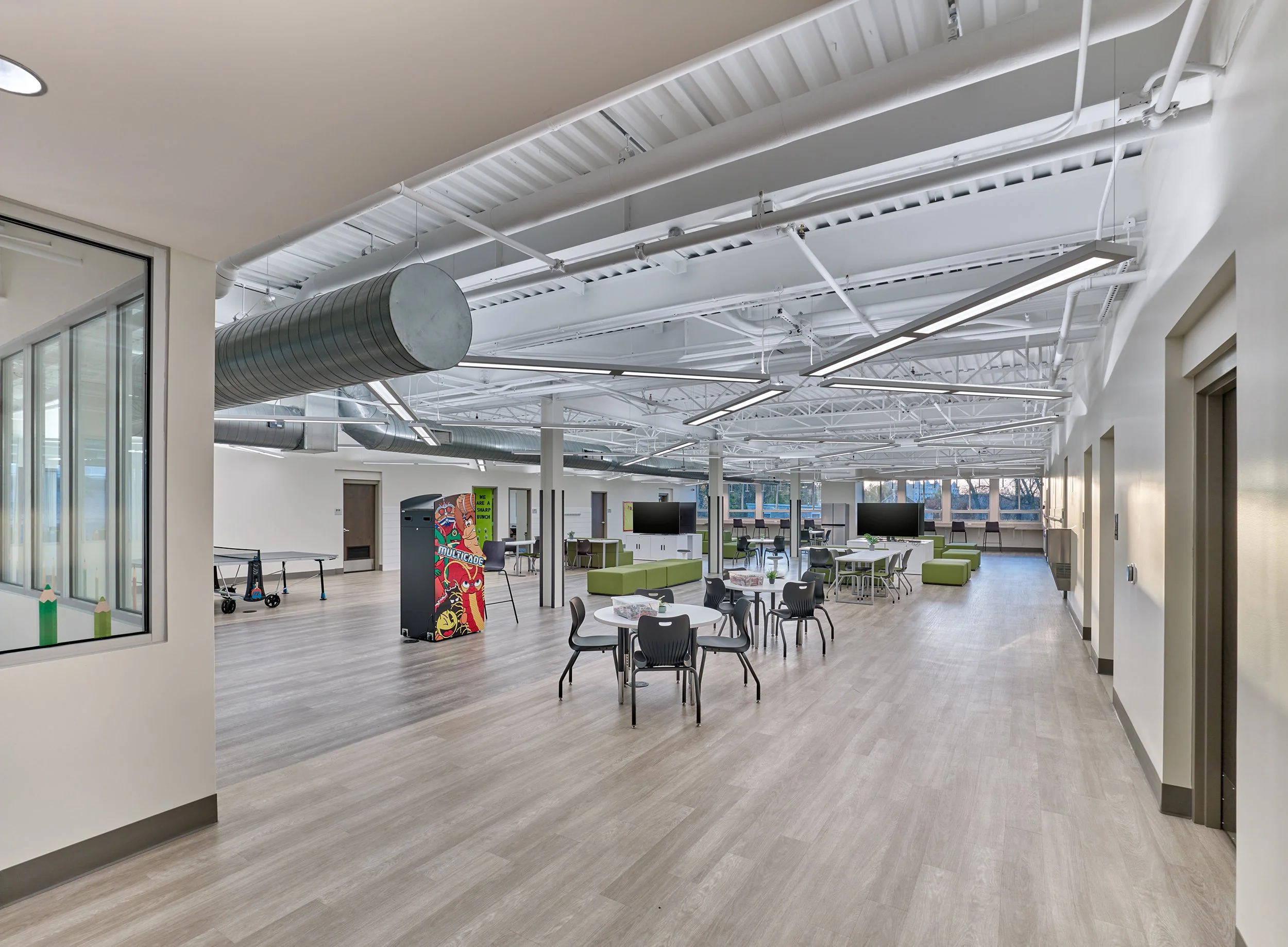 Open club community space with green seating, tables, arcade game, a large television, and ample natural light.