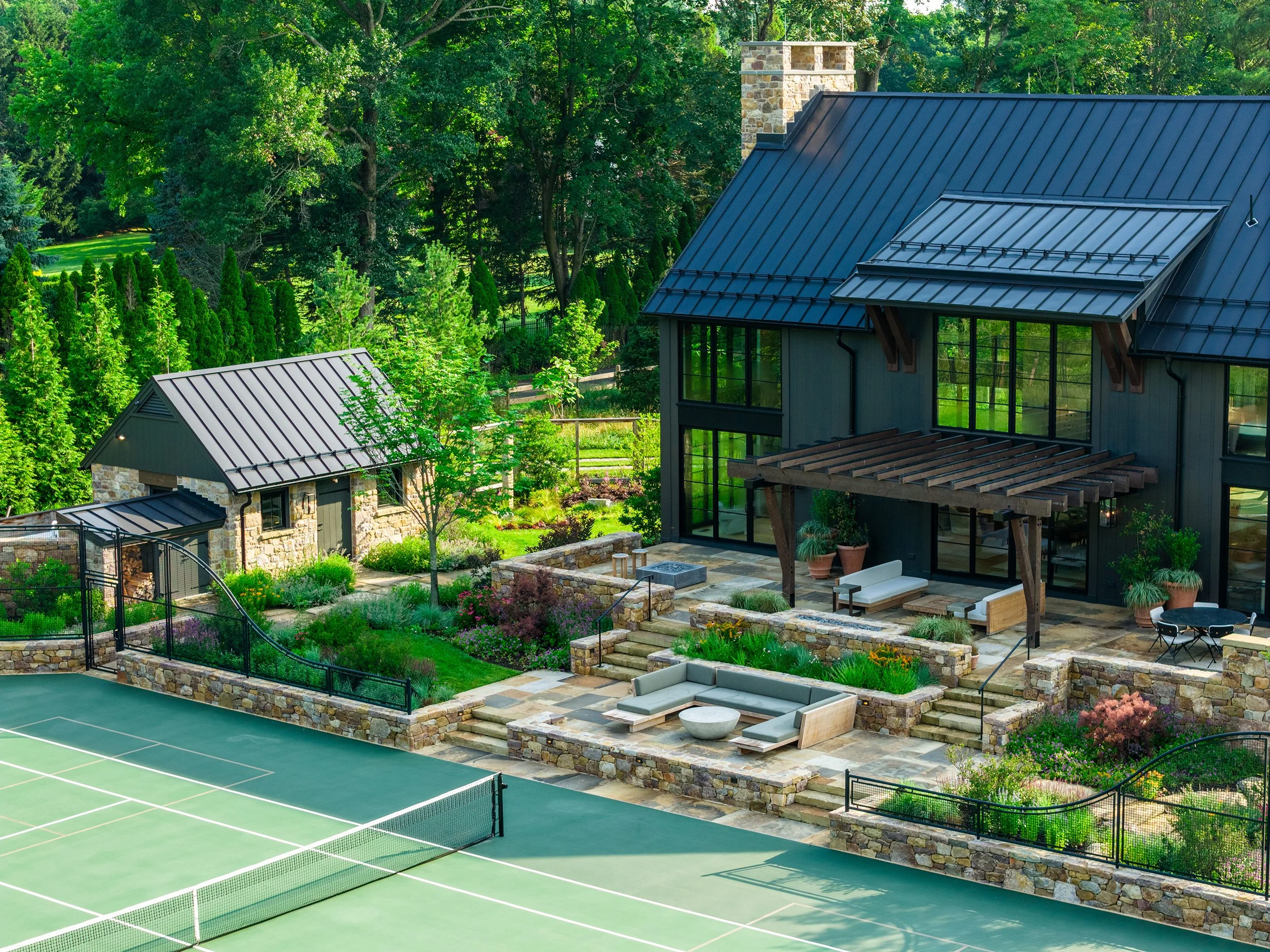 A luxurious outdoor backyard with a tennis court, stone patio, modern black barn, lush greenery, and landscaped garden.