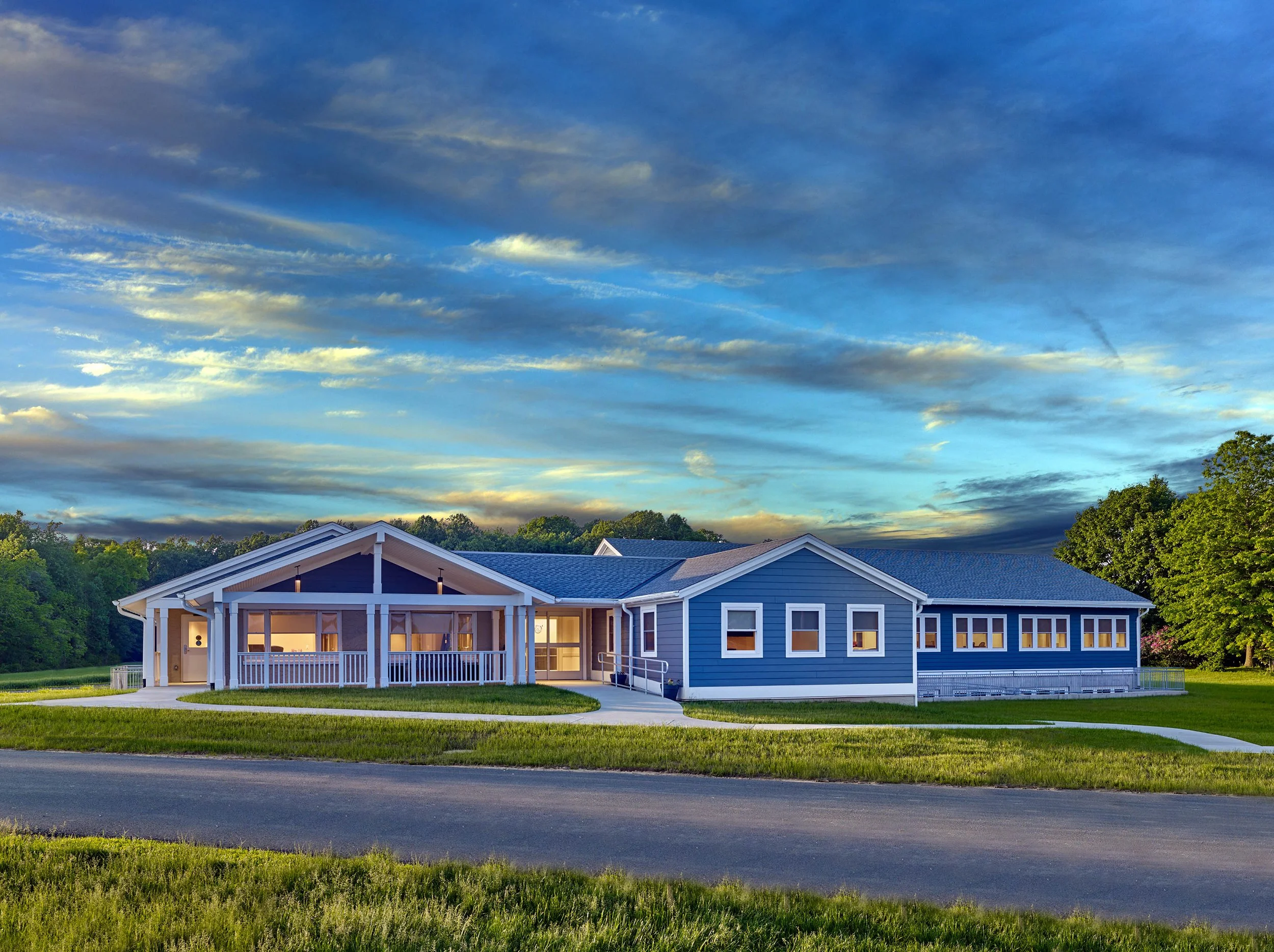 Modern blue house with large front porch and white trim, situated on a well-maintained lawn, with a paved walkway leading to the entrance, under a partly cloudy sky at dusk.