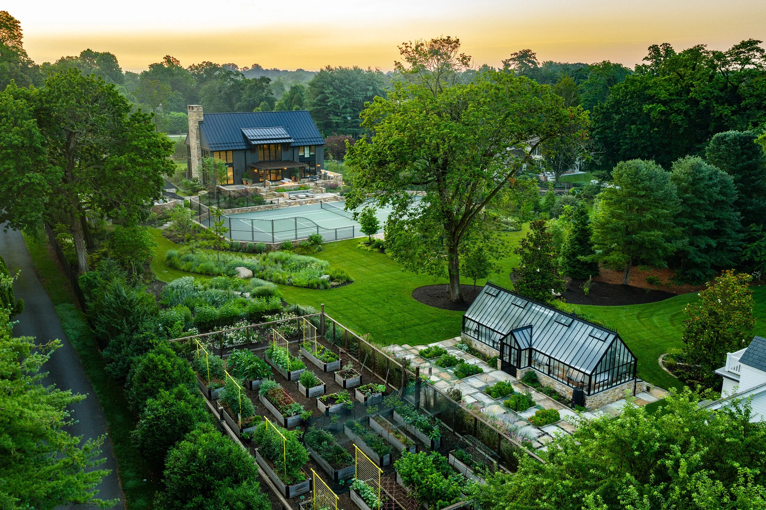A lush backyard with a garden, greenhouse, tennis court, and a modern barn, surrounded by tall trees and greenery.
