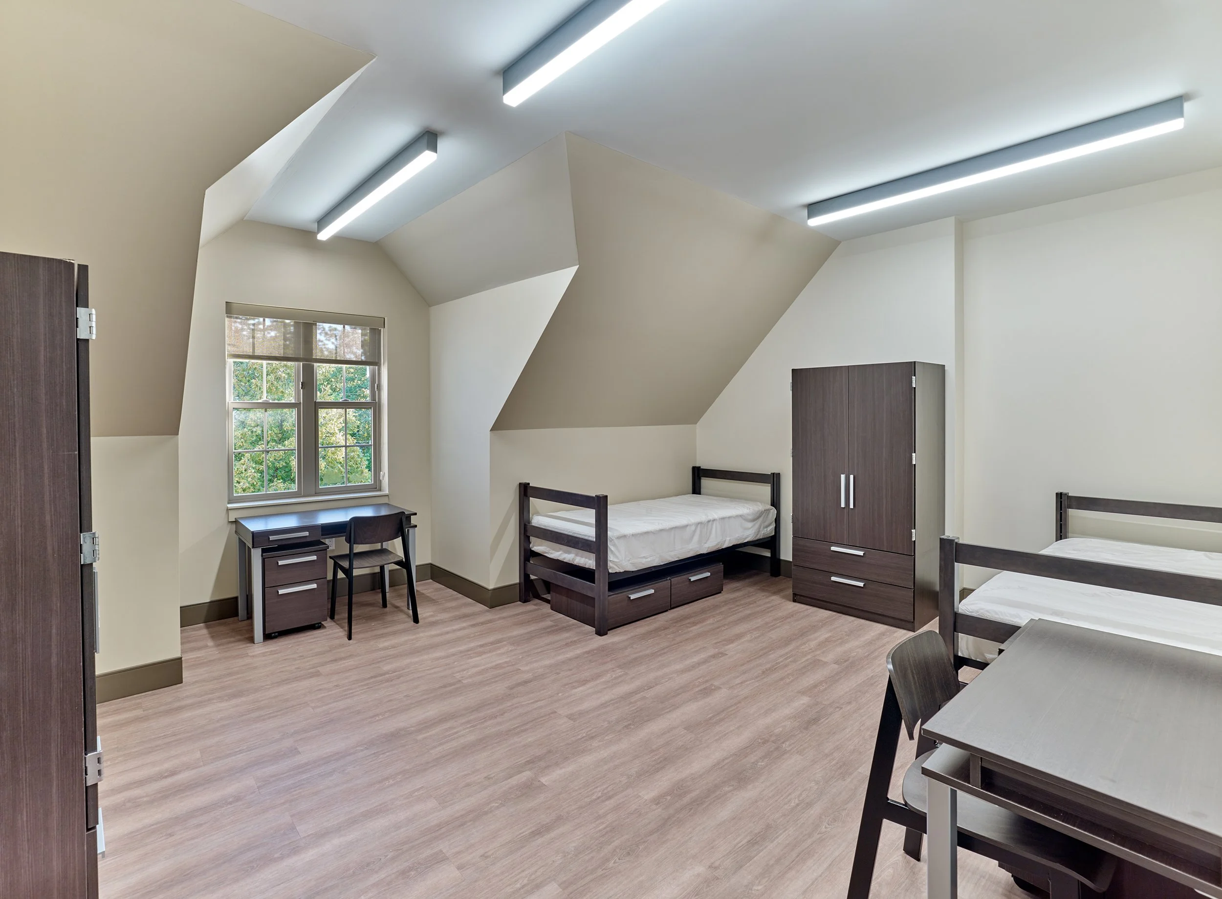 Dormitory room with two beds, a desk, a chair, a wardrobe, and wooden flooring.