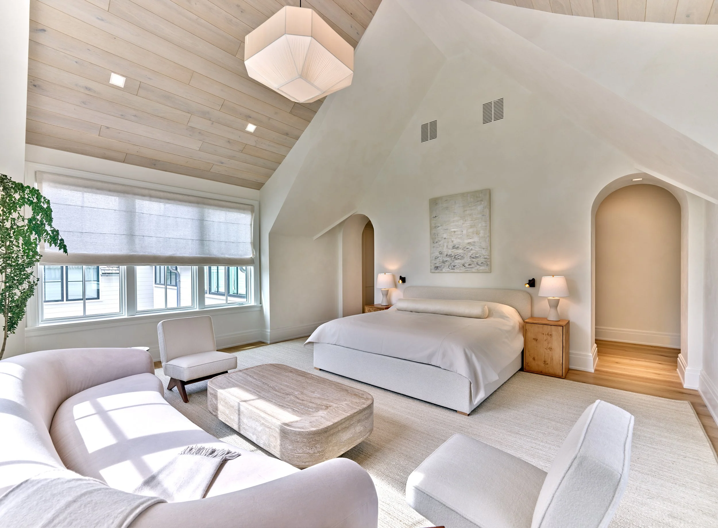 A spacious bedroom with a vaulted wood ceiling, large windows with curtains, a beige bed with a lamp on the side table, a cream-colored sofa, an armchair, a coffee table, and a potted plant.